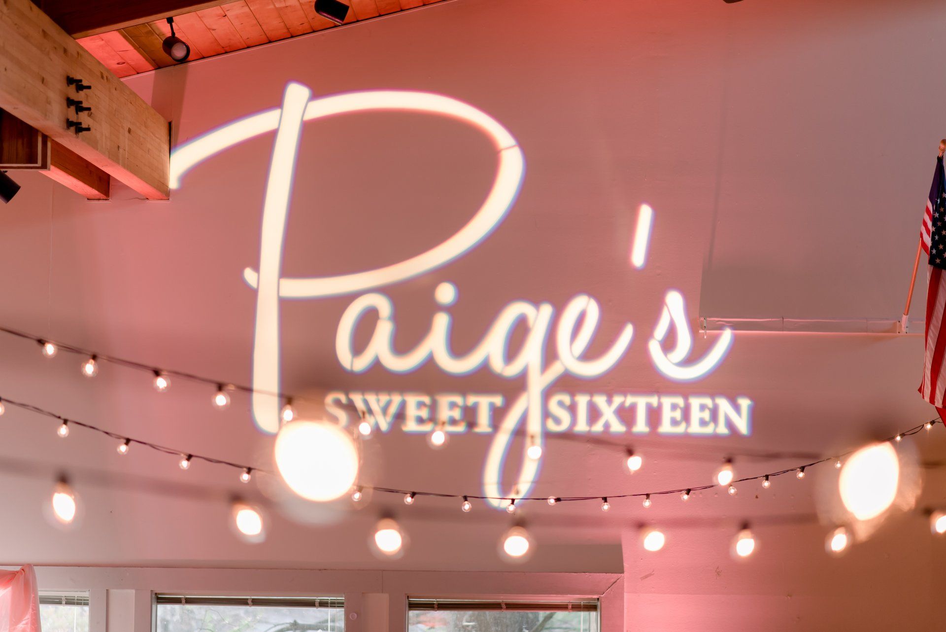 Paige’s Sweet 16 neon sign with string lights overhead in a pink-lit room