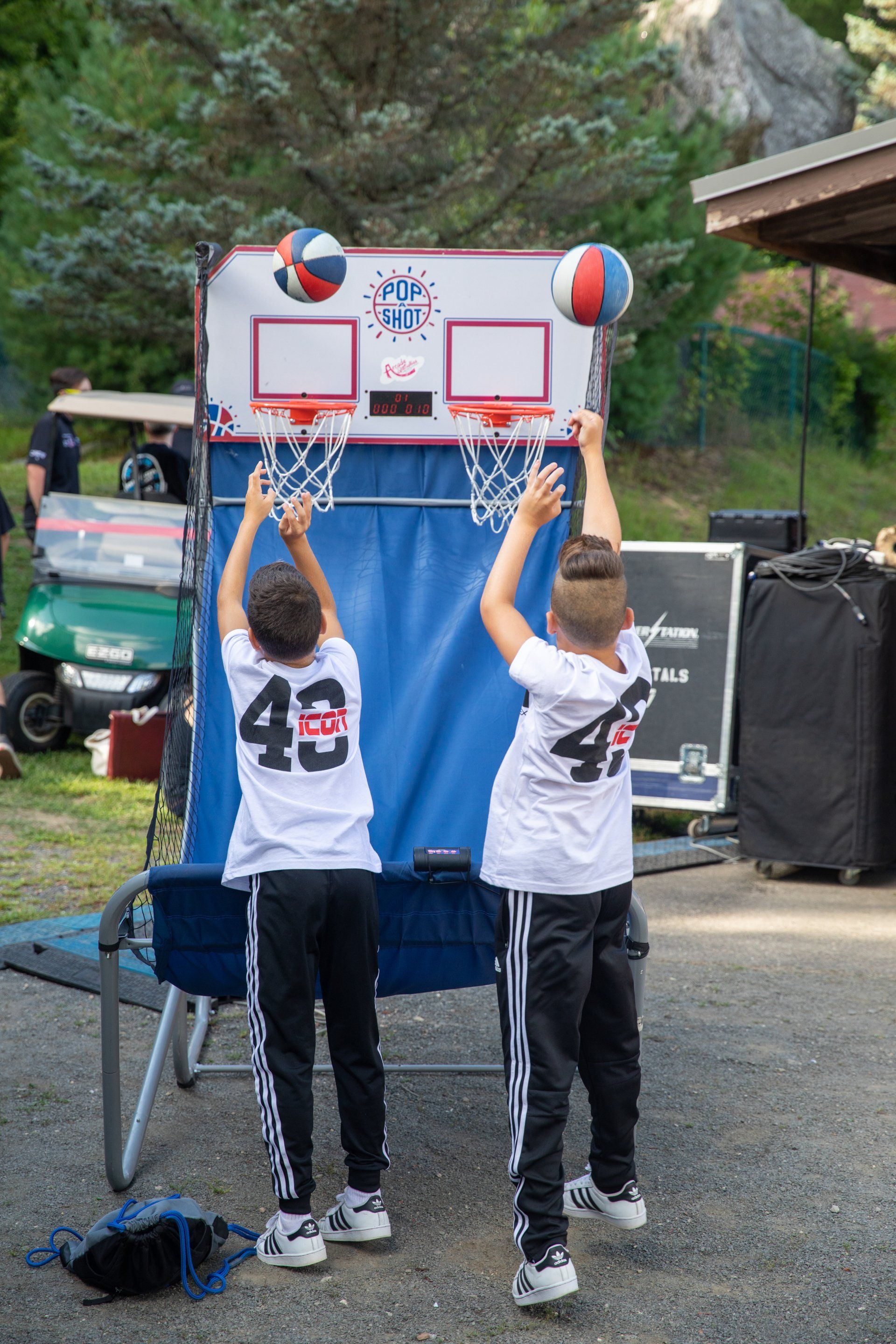 Two people in white jerseys shoot basketballs at a small outdoor hoop.