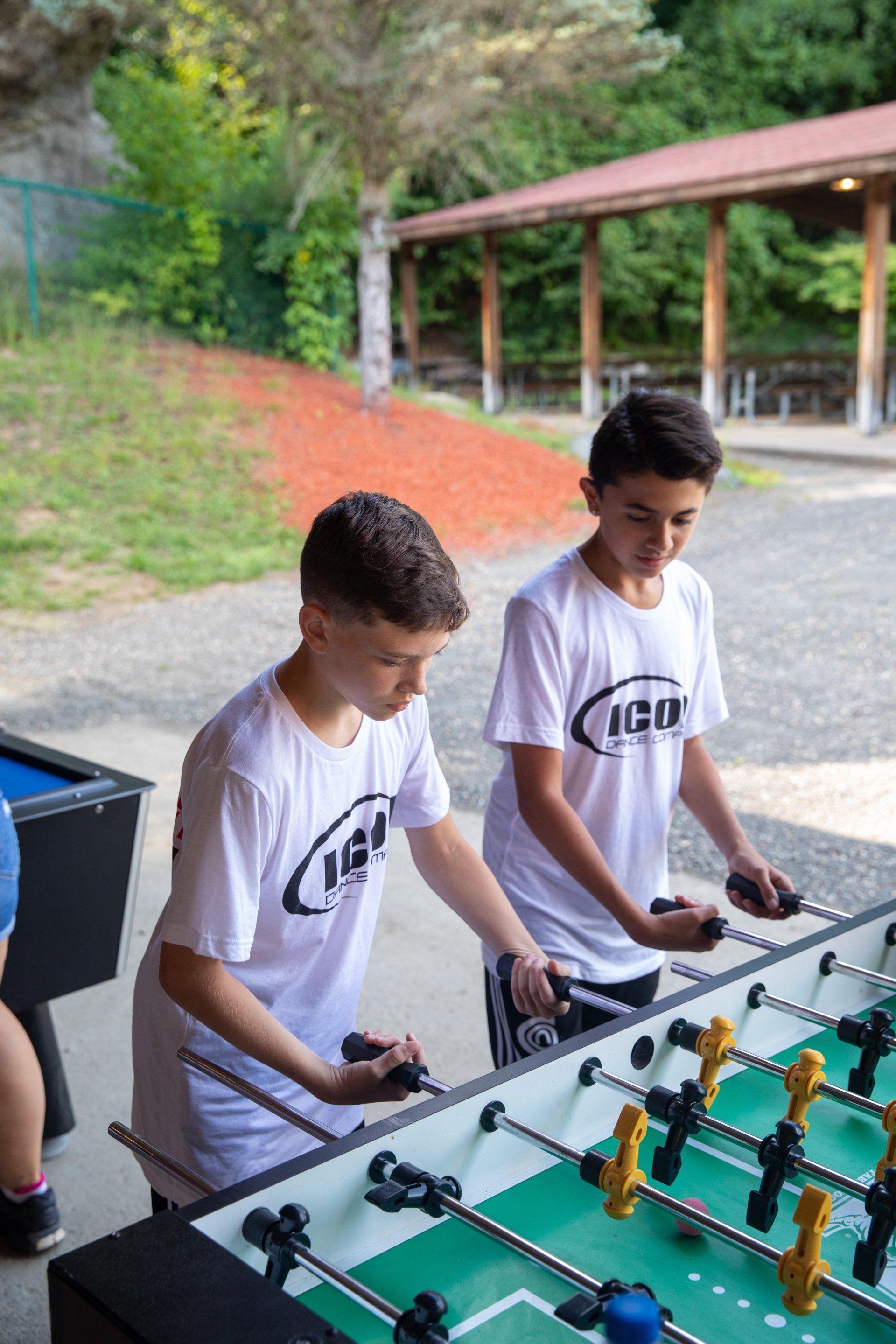 Two boys playing foosball outdoors at a table under a covered walkway.