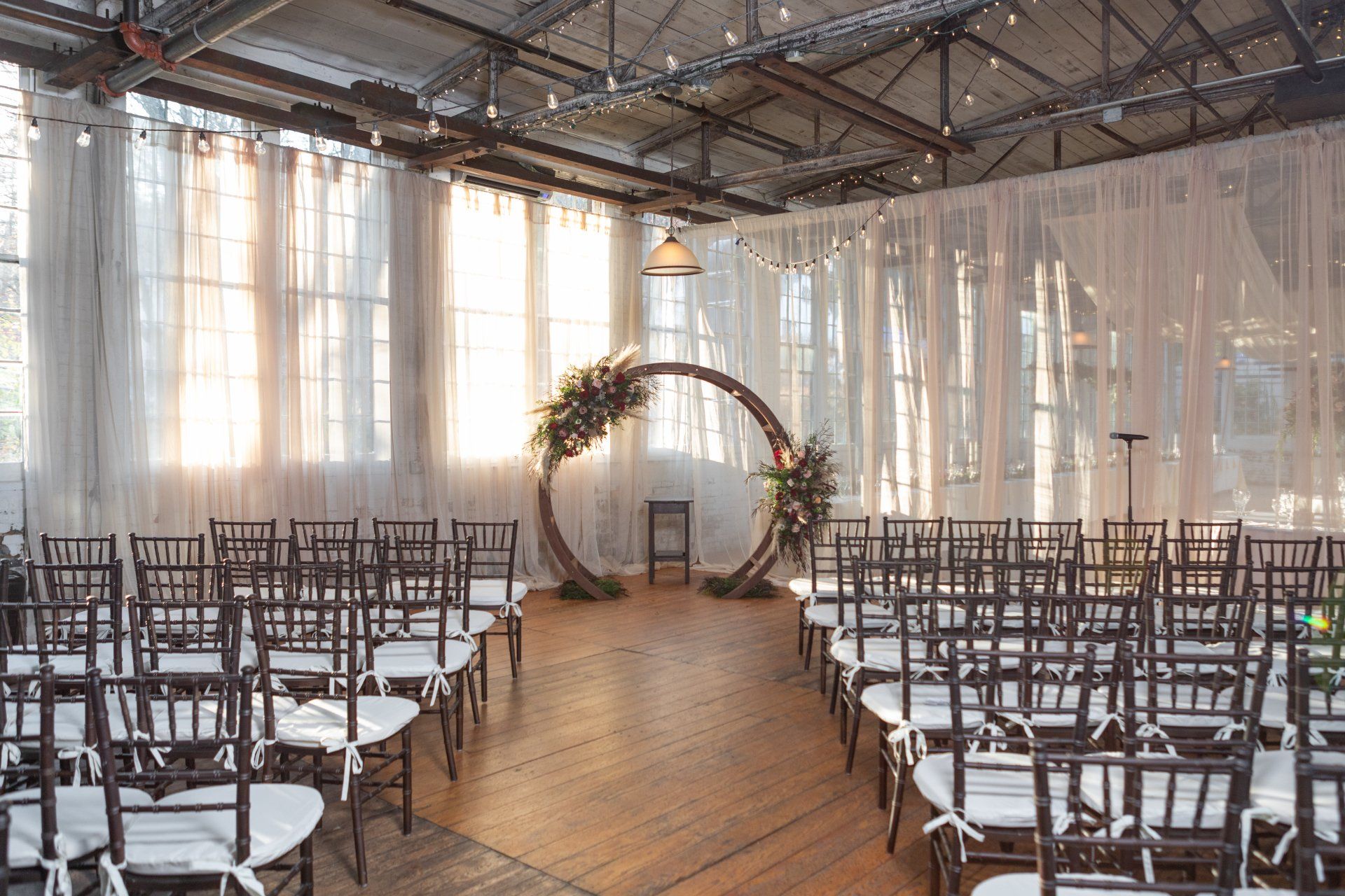 Rustic wedding ceremony setup with rows of chairs, wooden aisle, and floral arch in a bright hall