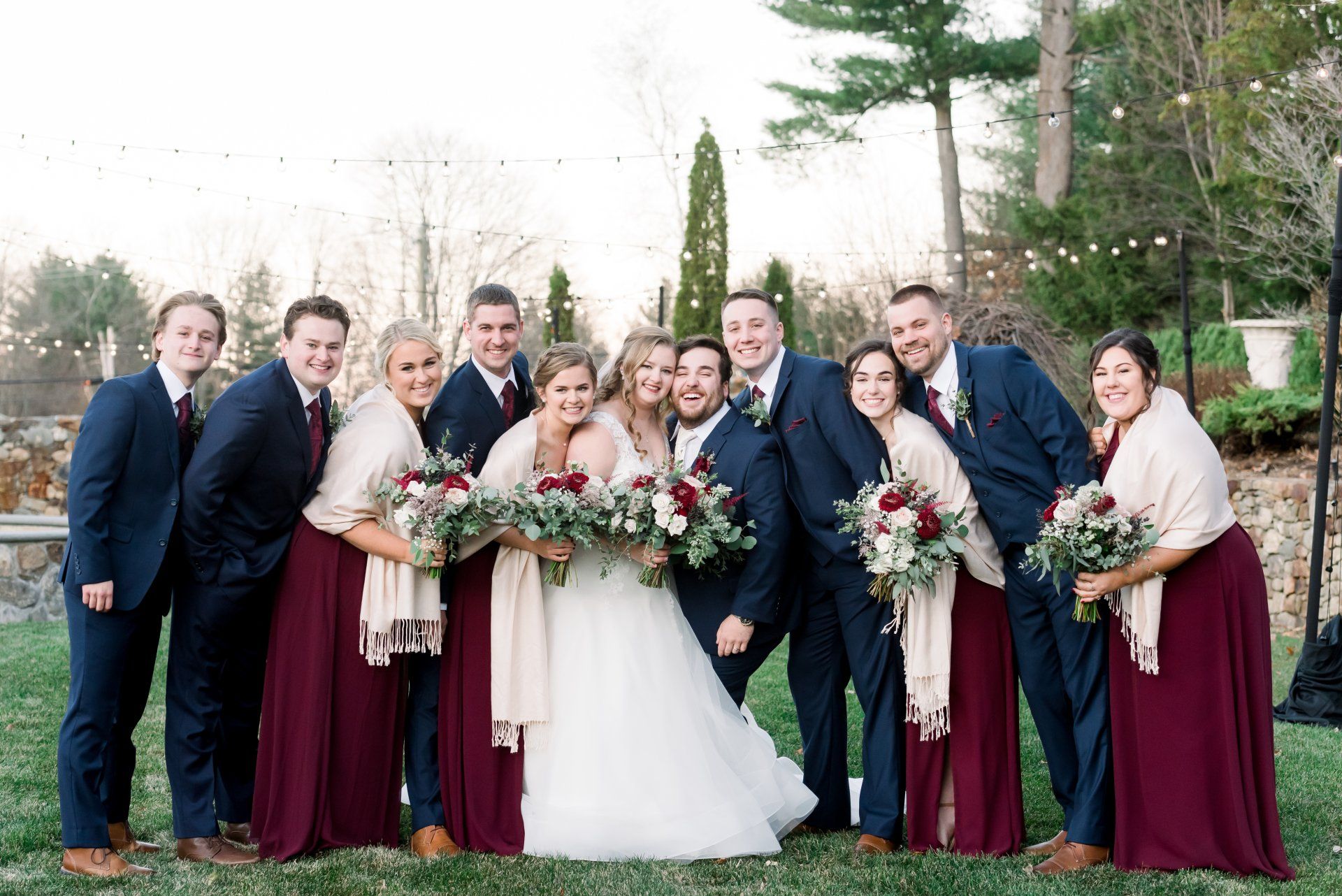 Wedding party posing outdoors in burgundy dresses and dark suits, holding bouquets around the bride and groom.