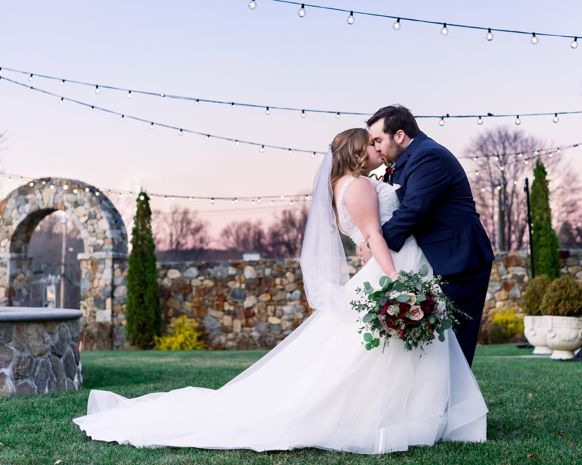 Bride and groom kissing outdoors at sunset, bride in white gown holding a bouquet