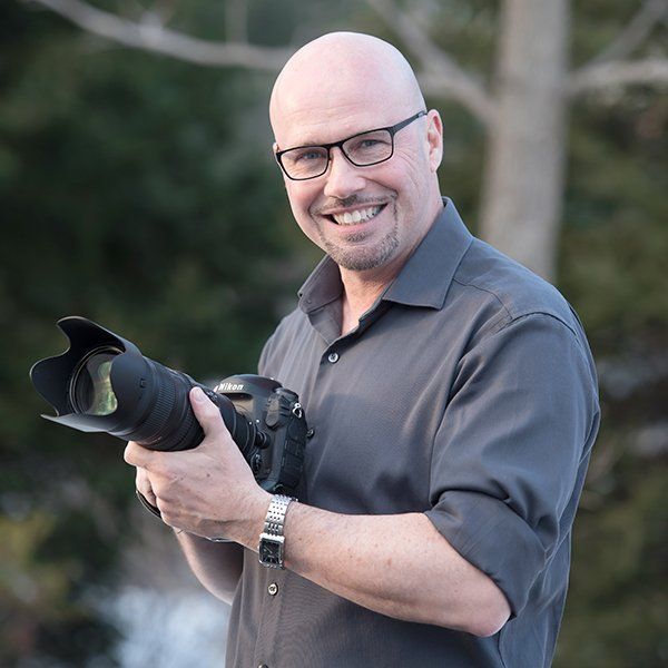 Smiling person holding a camera with a telephoto lens outdoors, wearing a gray shirt and glasses.