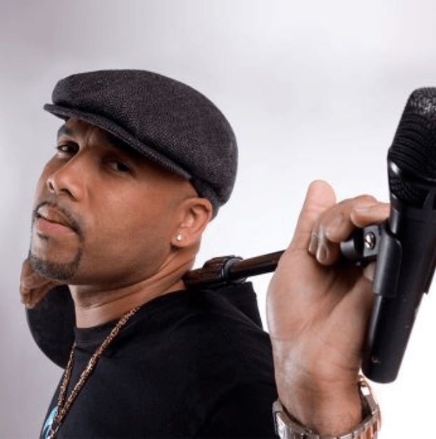 Person in a dark flat cap holding a microphone over their shoulder, wearing a black shirt and chain.