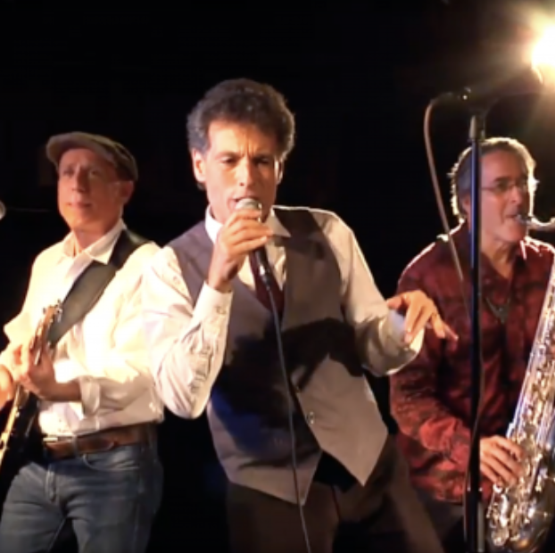 Three musicians performing on stage, with a singer holding a microphone and a saxophone player beside him.