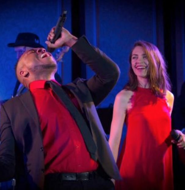 Singer in red shirt and hat drinks from a bottle while a woman in a red dress smiles onstage under blue lights