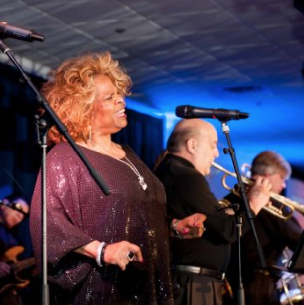 Three musicians performing onstage under blue lighting, with microphones and a smiling singer in the foreground