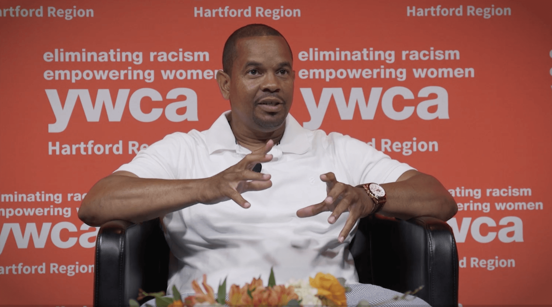 Man speaking at a YMCA Hartford Region event, gesturing with hands before a red branded backdrop
