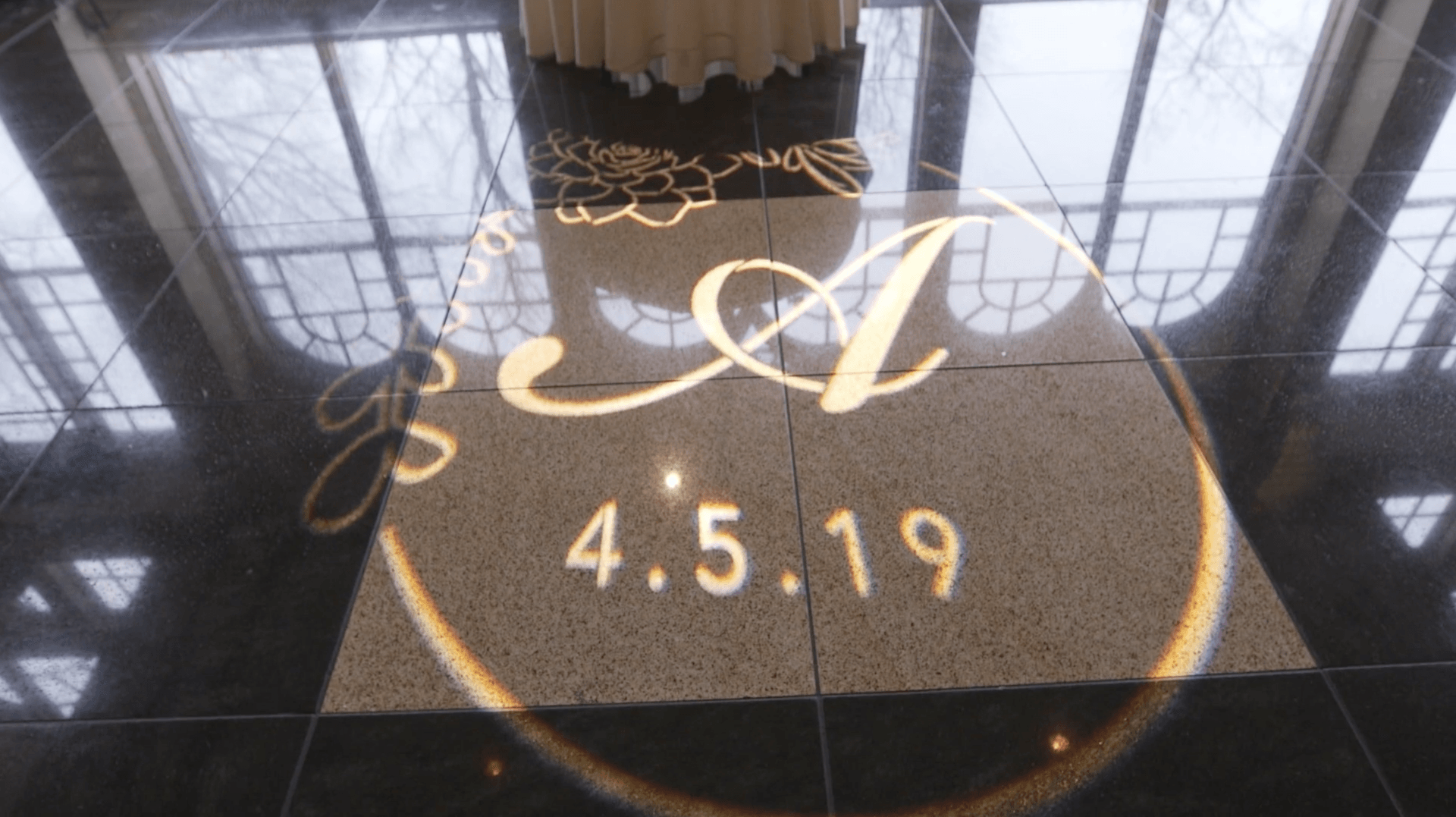 Gold script monogram “A” and date “4.5.19” projected on a glossy black floor.