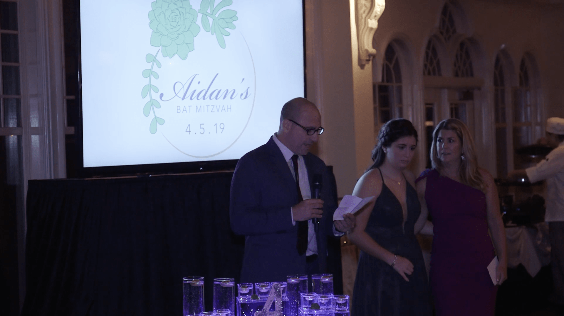 Three people in formal wear stand at a reception beside a lit screen and drink table