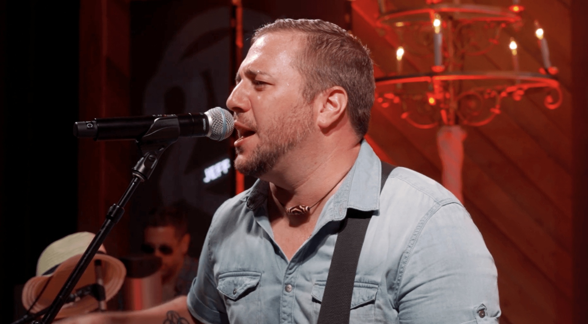 Singer performing into a microphone on a red-lit stage, wearing a light blue shirt and guitar strap