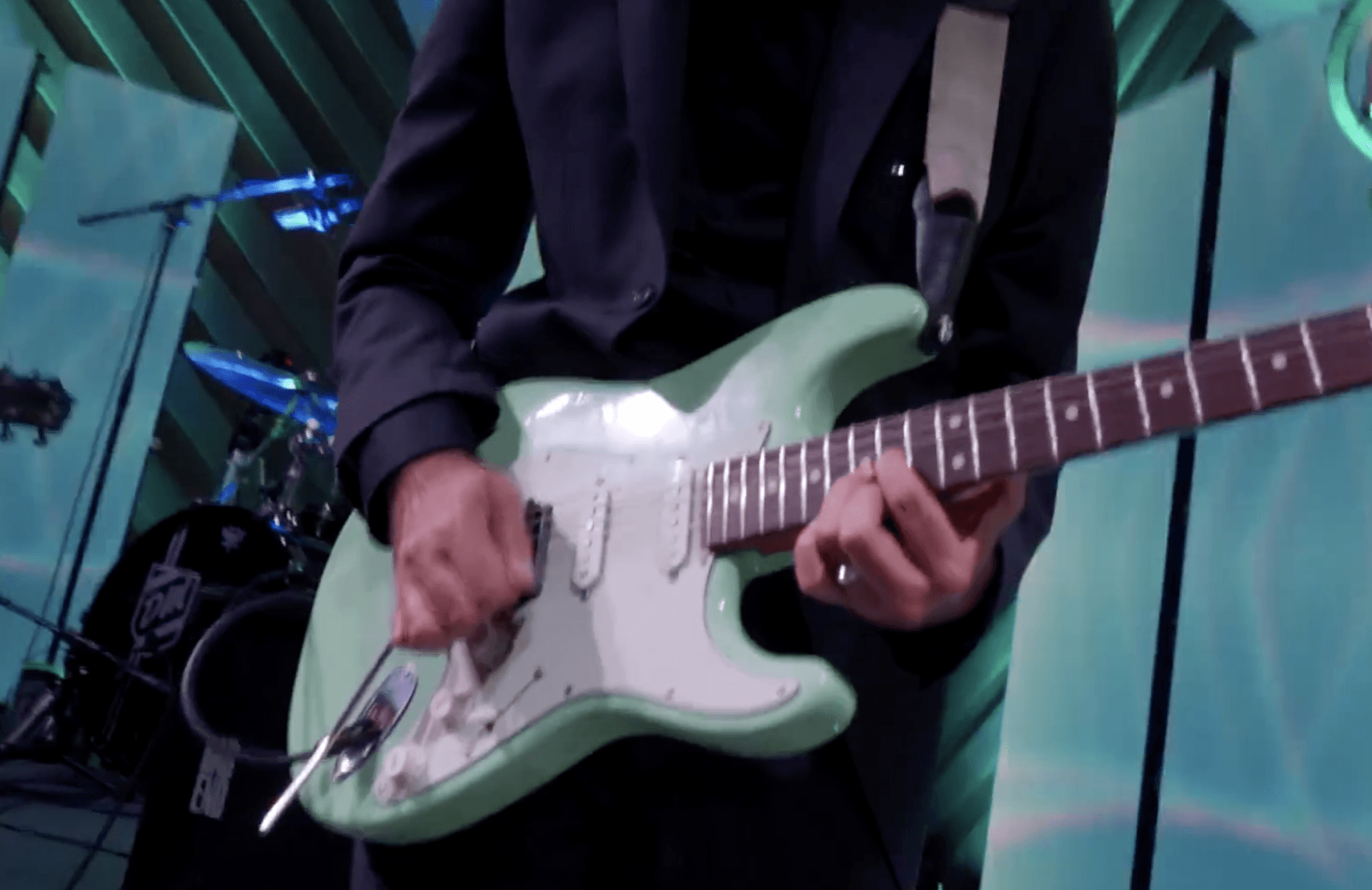 Musician playing a mint-green electric guitar on stage with drums in the background