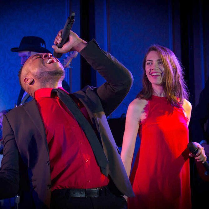 Singer in red shirt and woman in red dress performing on stage under blue and purple lights