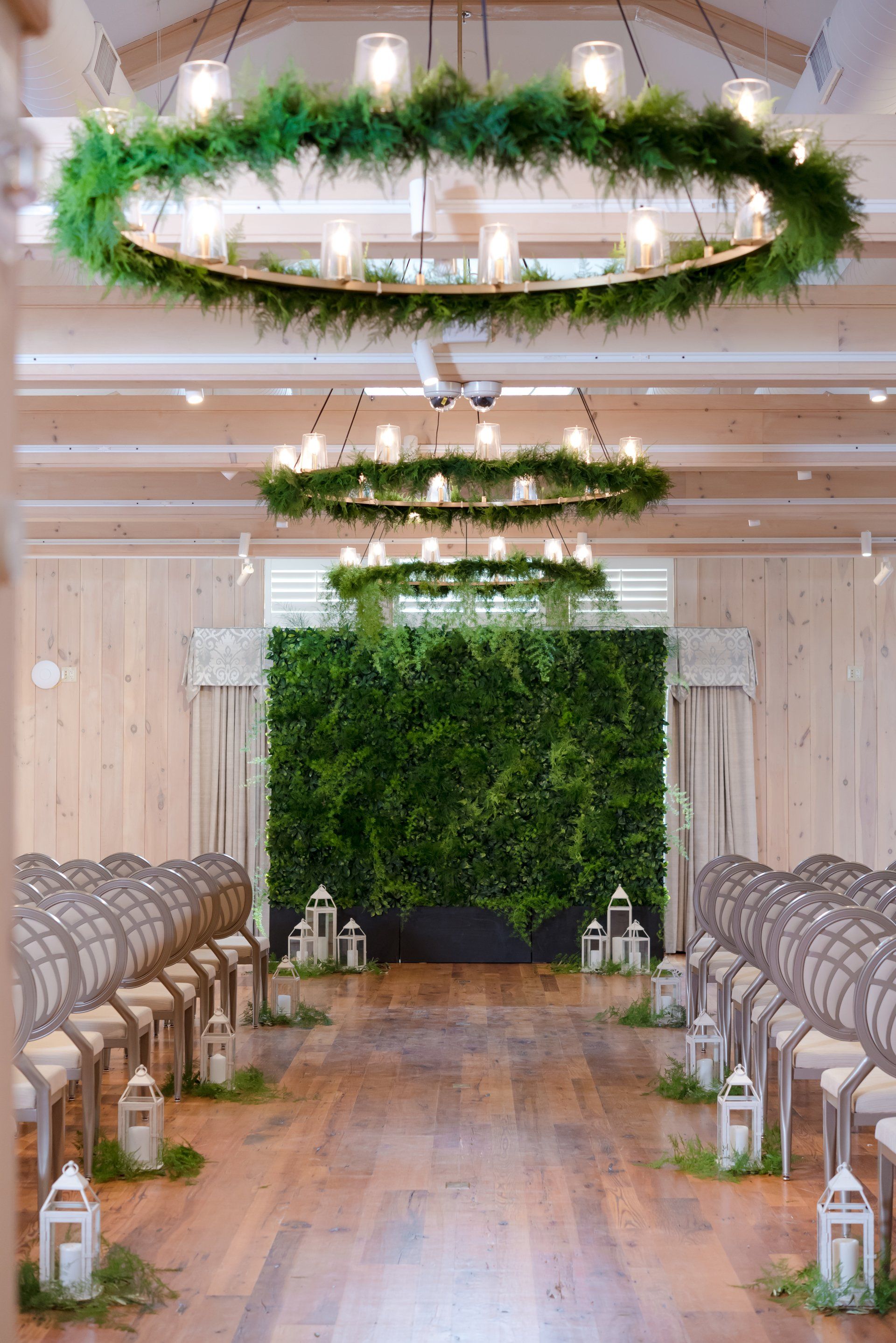 A decorated wedding aisle with wooden chairs, lanterns, greenery, and hanging floral wreaths in a rustic venue