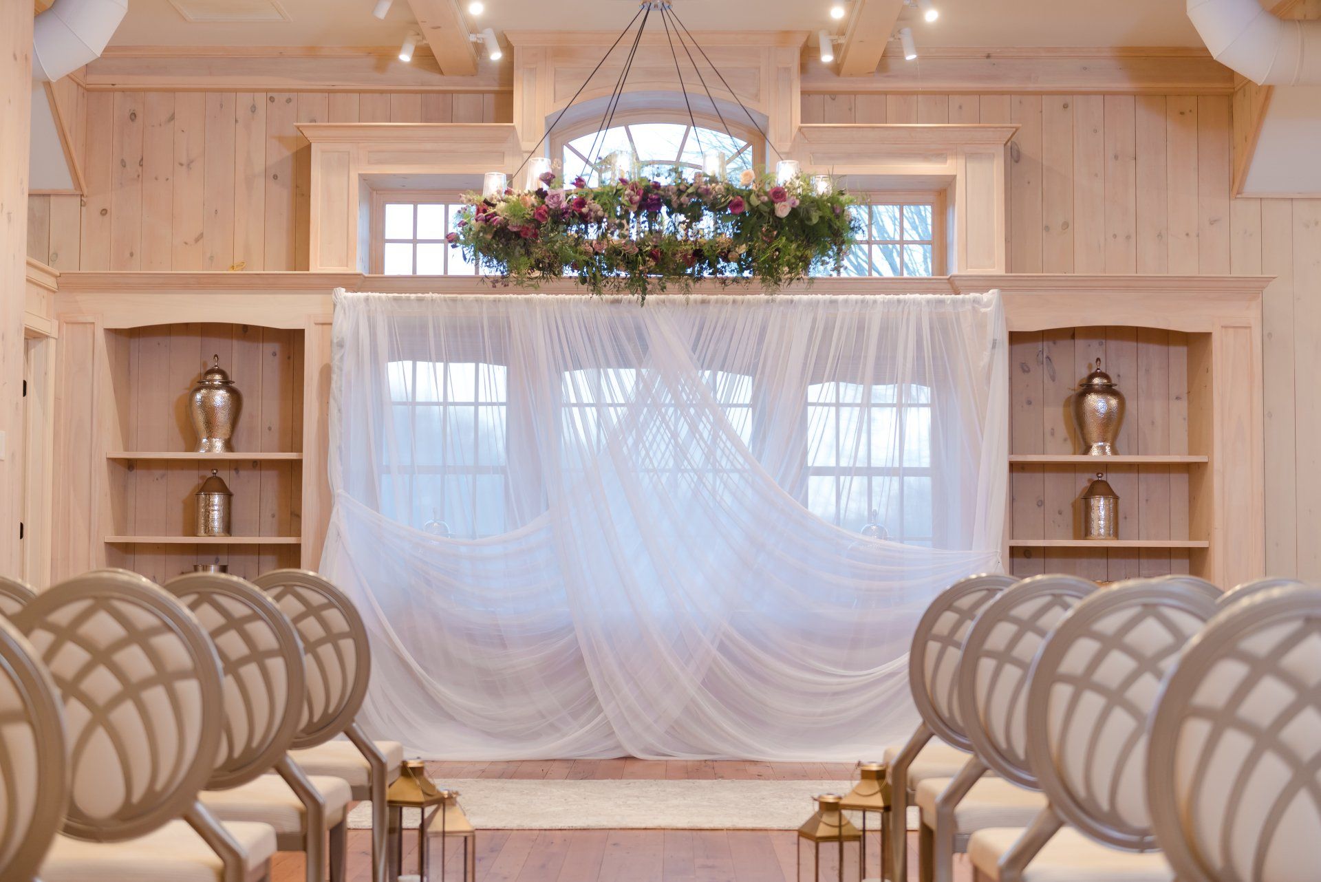 Softly draped wedding backdrop with sheer white curtains, floral chandelier, and rows of chairs in a bright wood chapel