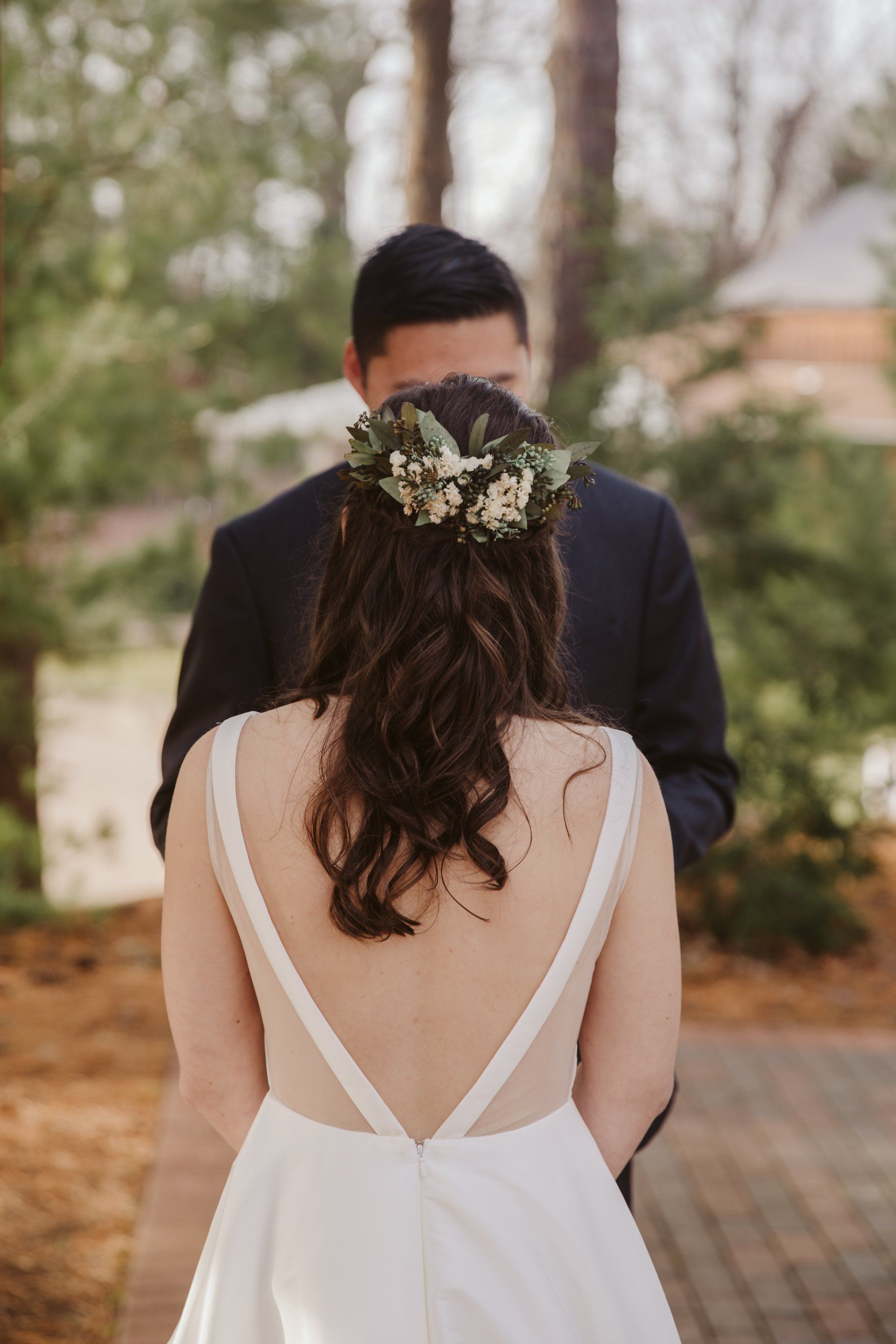 Bride in white dress facing groom in dark suit, outdoors with blurred trees and a floral crown.
