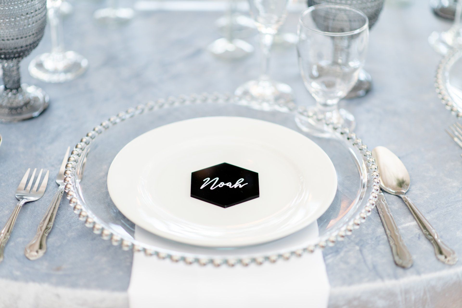 Elegant place setting with white plates, crystal chargers, and a black napkin card on a silver tablecloth