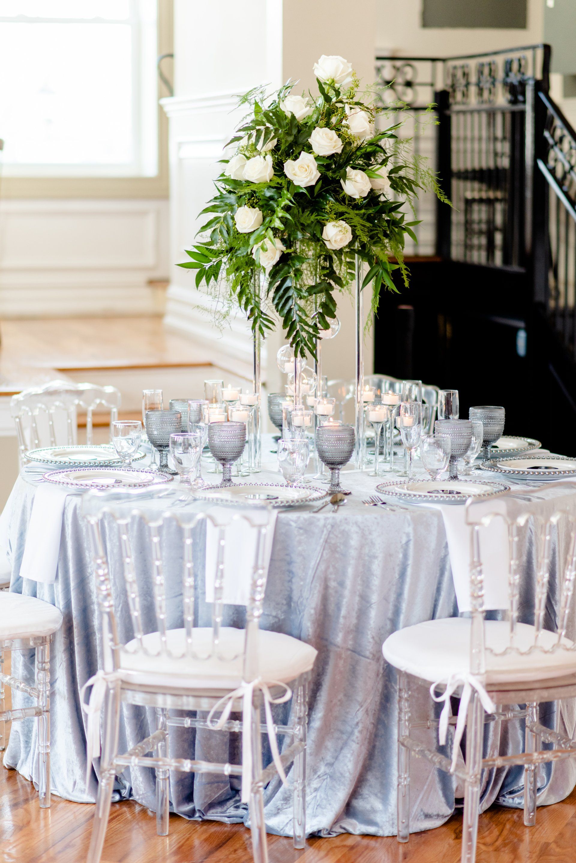 Round banquet table with silver tablecloth, glassware, and tall white floral centerpiece in a bright room