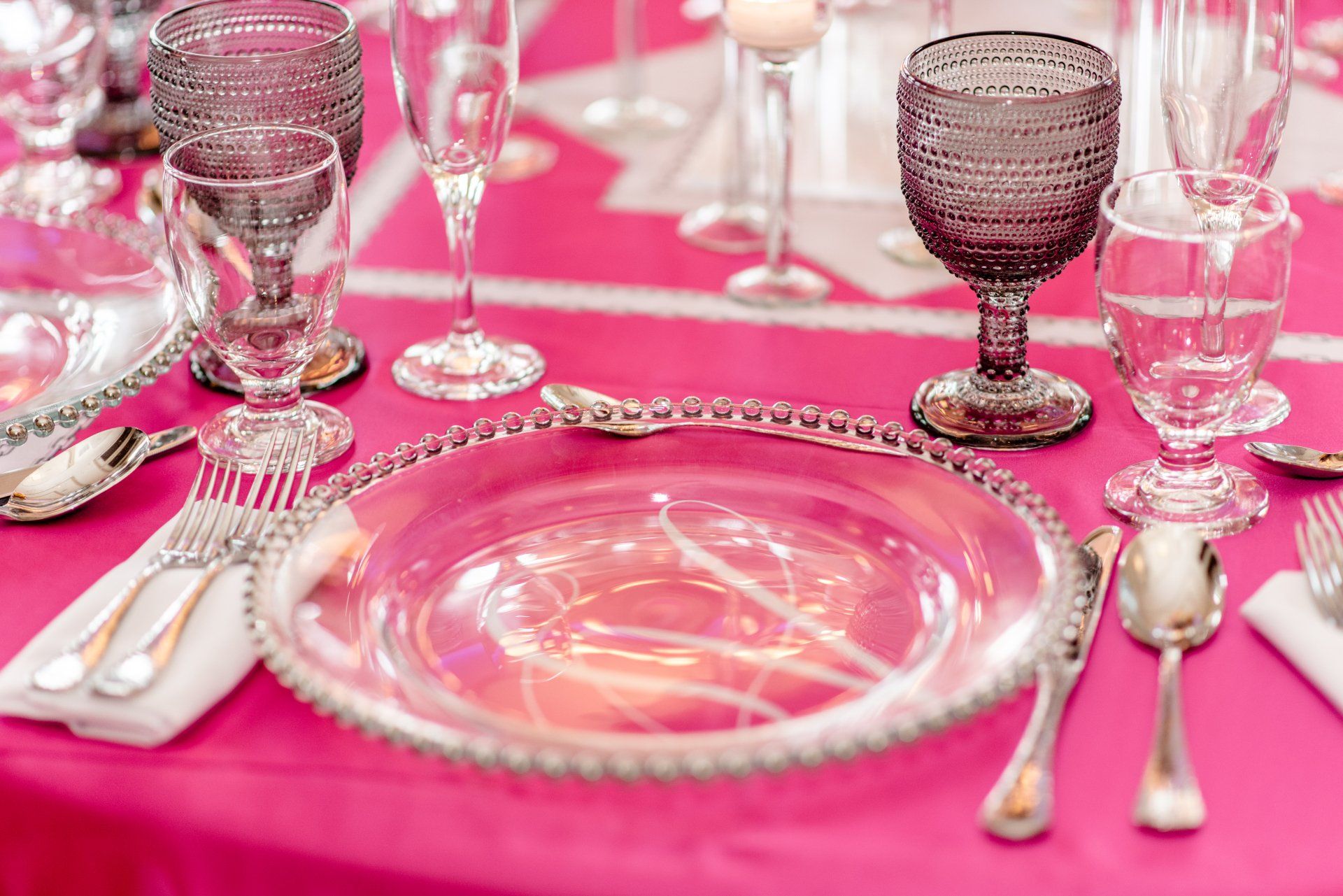 Pink table setting with clear glass plates, silverware, and decorative goblets on a bright pink tablecloth
