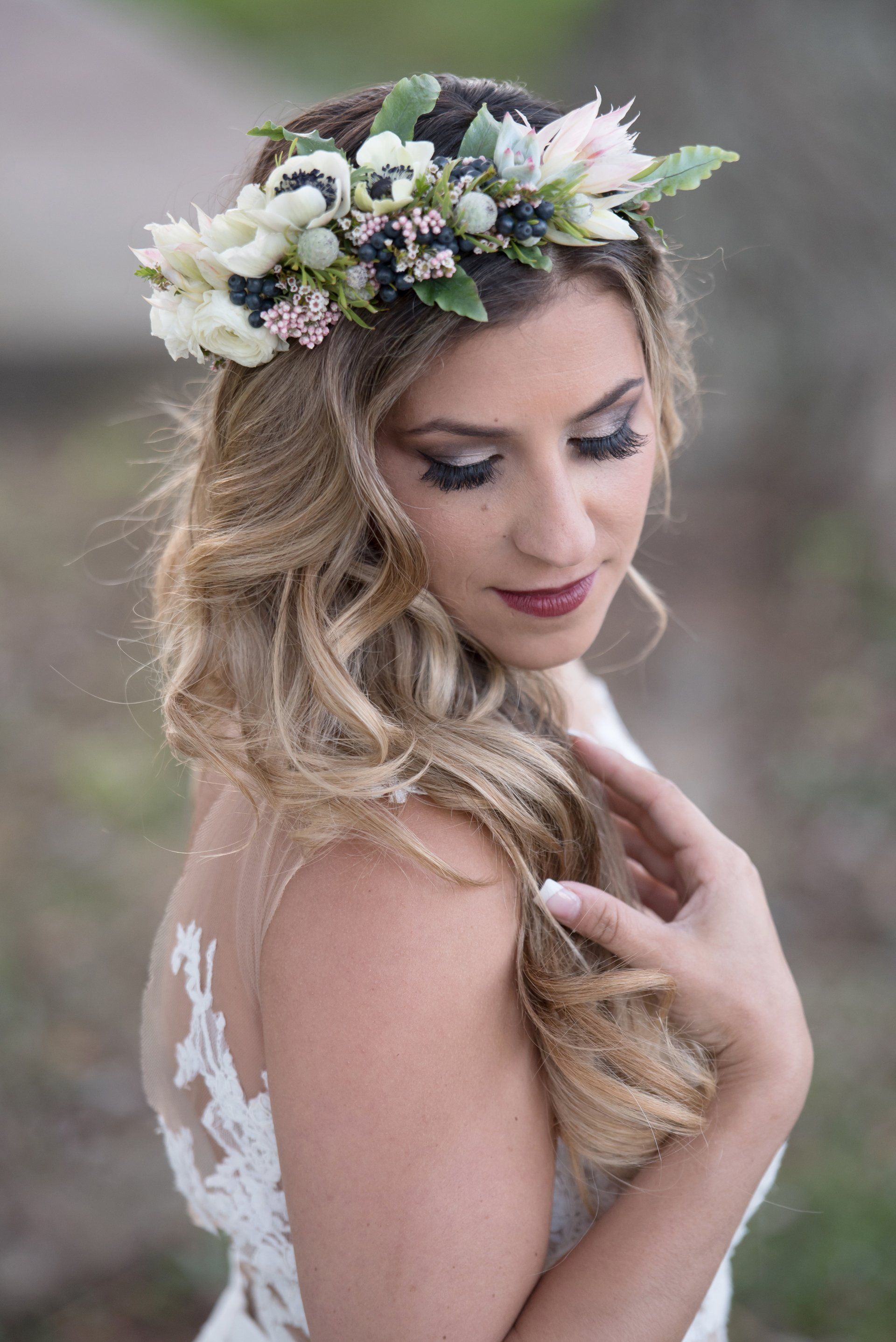Woman in a floral crown and white lace dress, gazing downward in a soft, natural outdoor setting