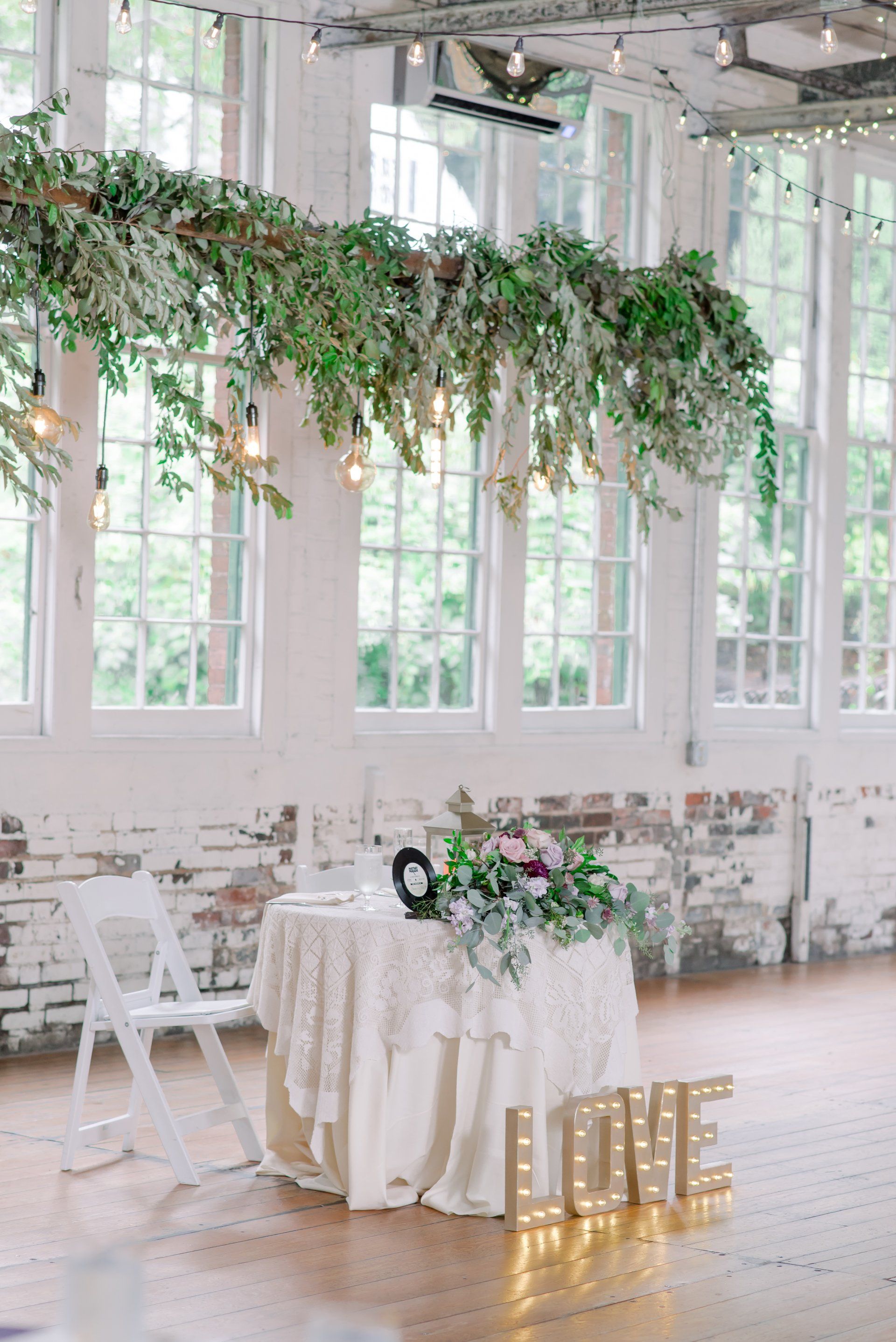 Bright white wedding reception setup with greenery, floral table, and gold “LOVE” letters by tall windows