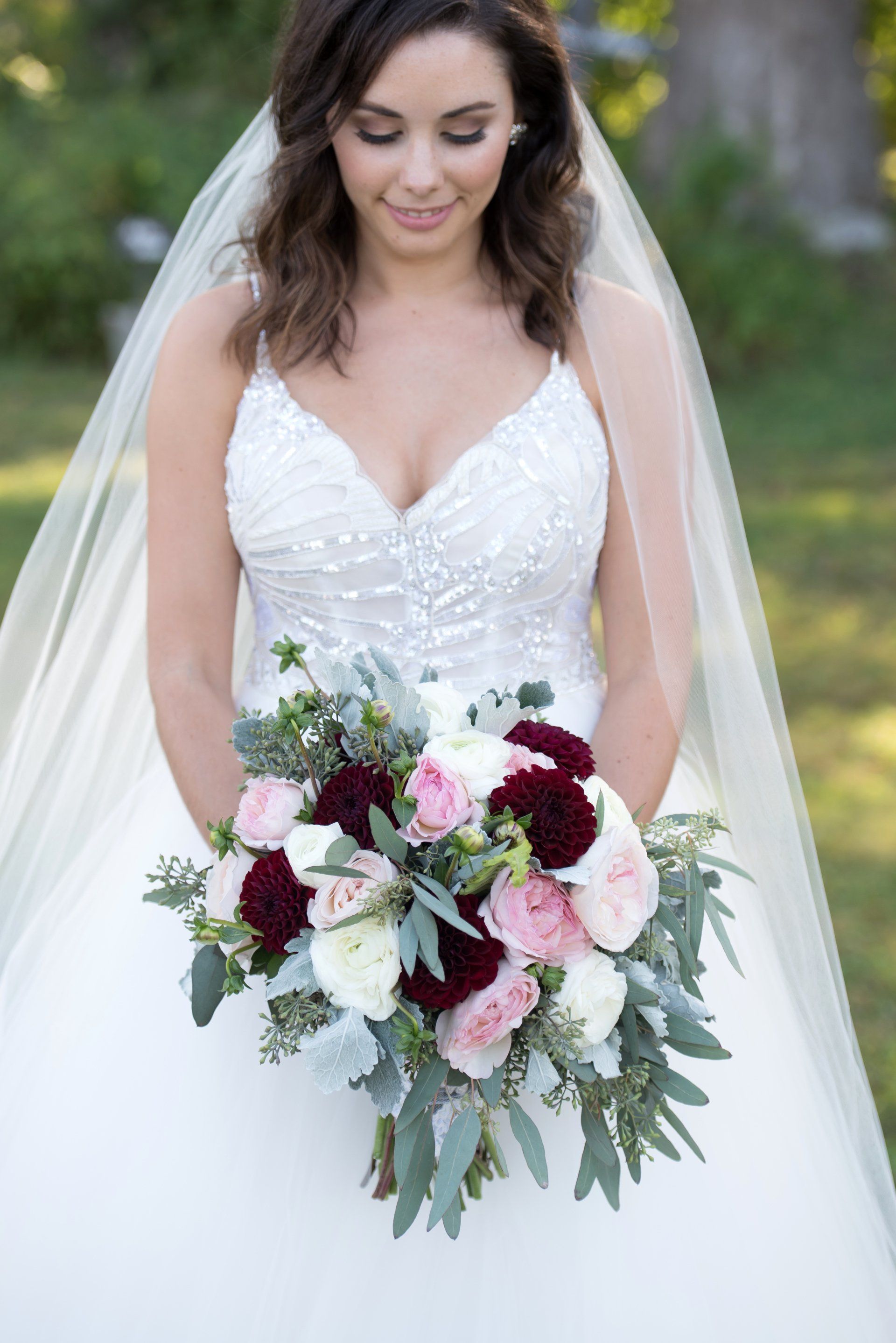 Bride in a white gown holding a bouquet of burgundy, pink, and white flowers outdoors