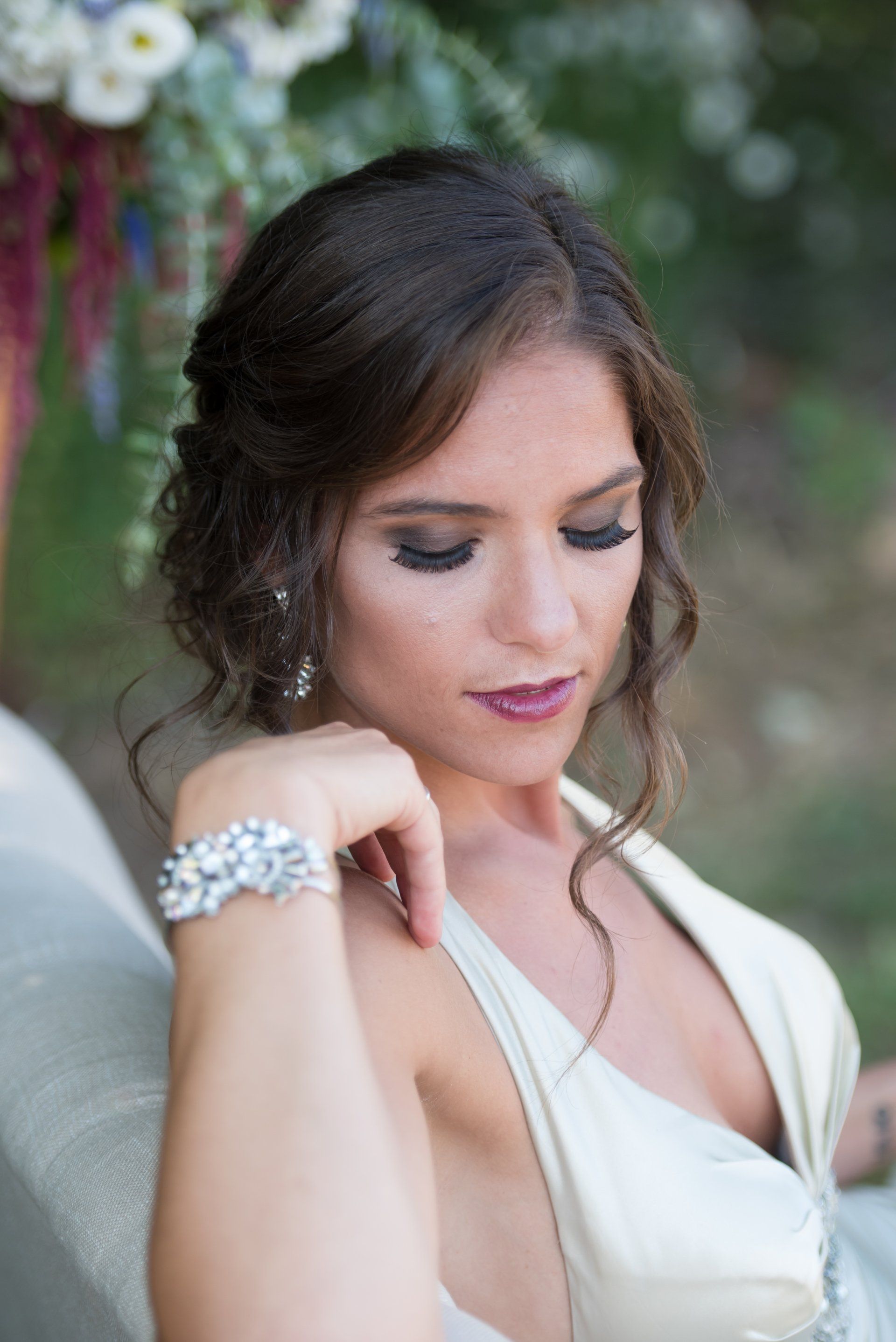 Woman in a white dress looking down, wearing a silver bracelet and necklace, against a blurred floral background