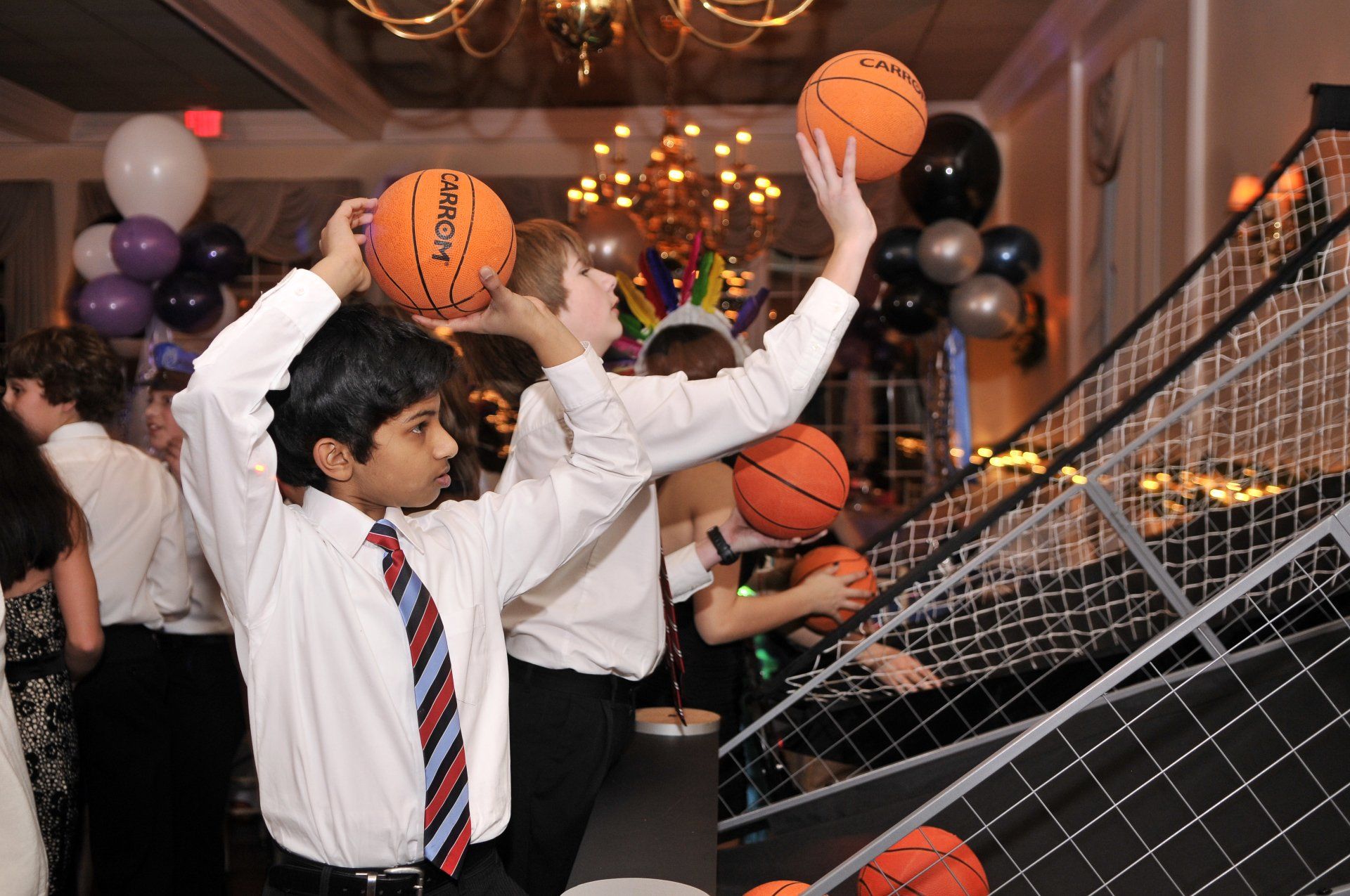 People tossing basketballs into a hoop at an indoor party with balloons and decorations