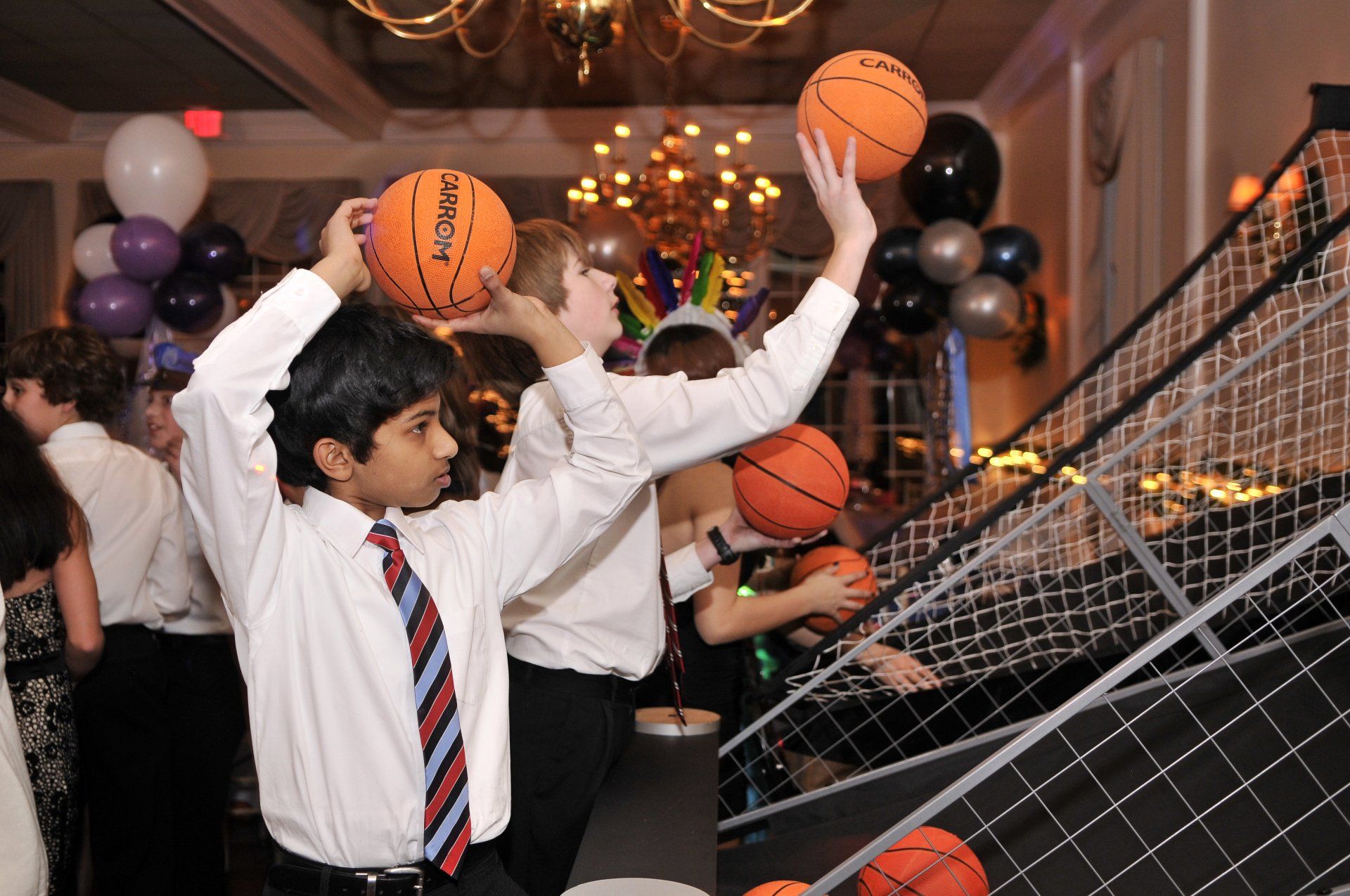 People tossing basketballs at an arcade game amid balloons and party lights