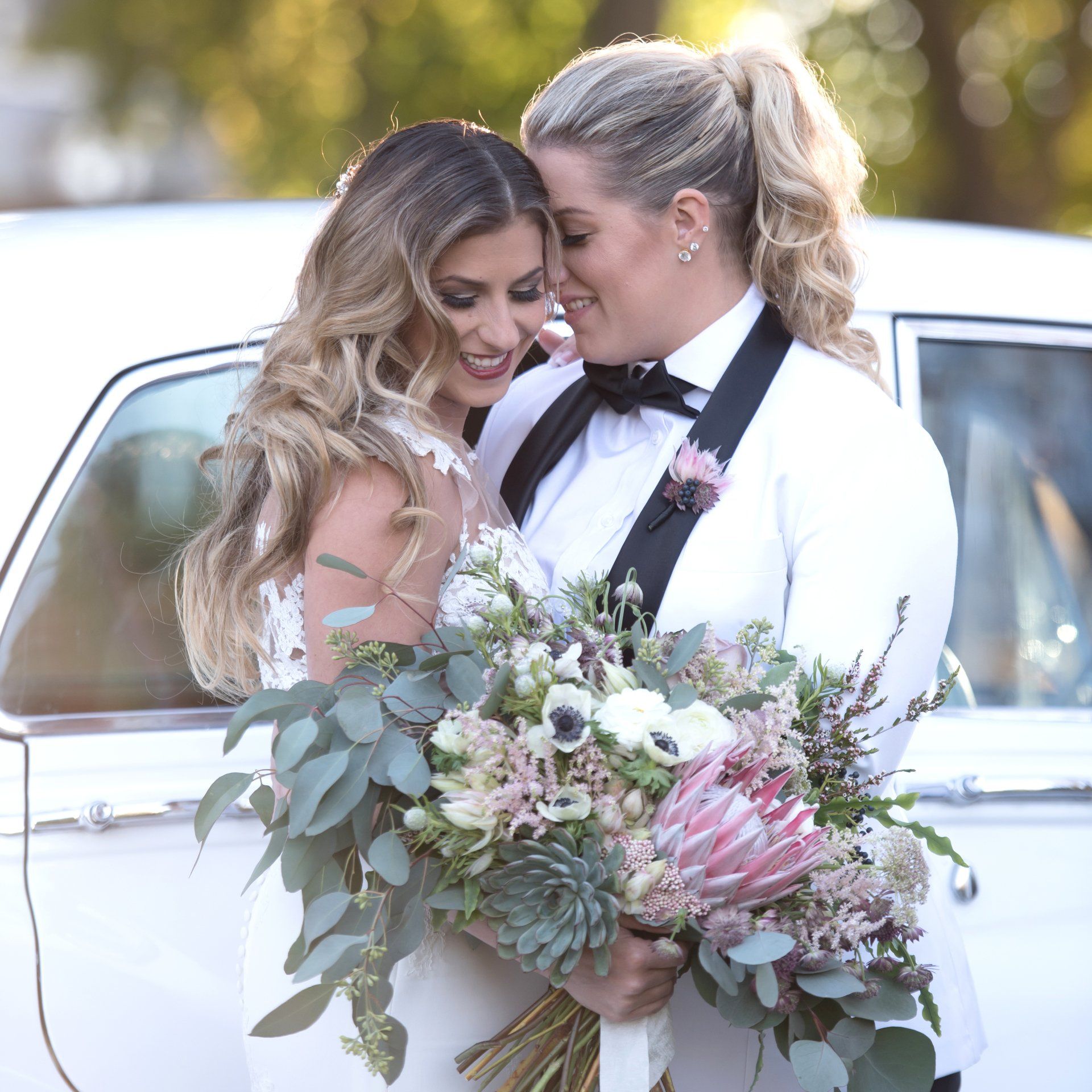 Two women in formal attire embrace beside a white car, holding a large bouquet of flowers.