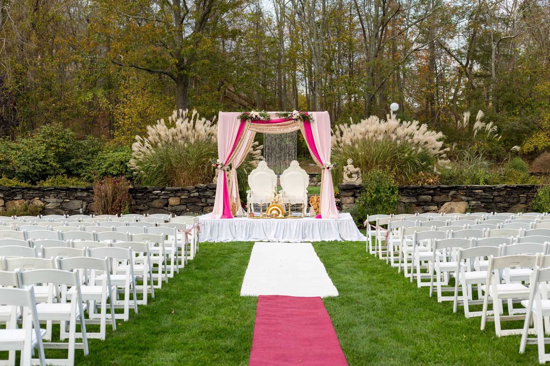 Outdoor wedding aisle with white chairs, pink runner, and floral arch in a garden setting