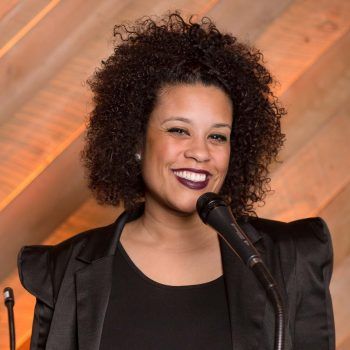 Woman smiling at a microphone onstage, wearing a black top against a warm wooden backdrop