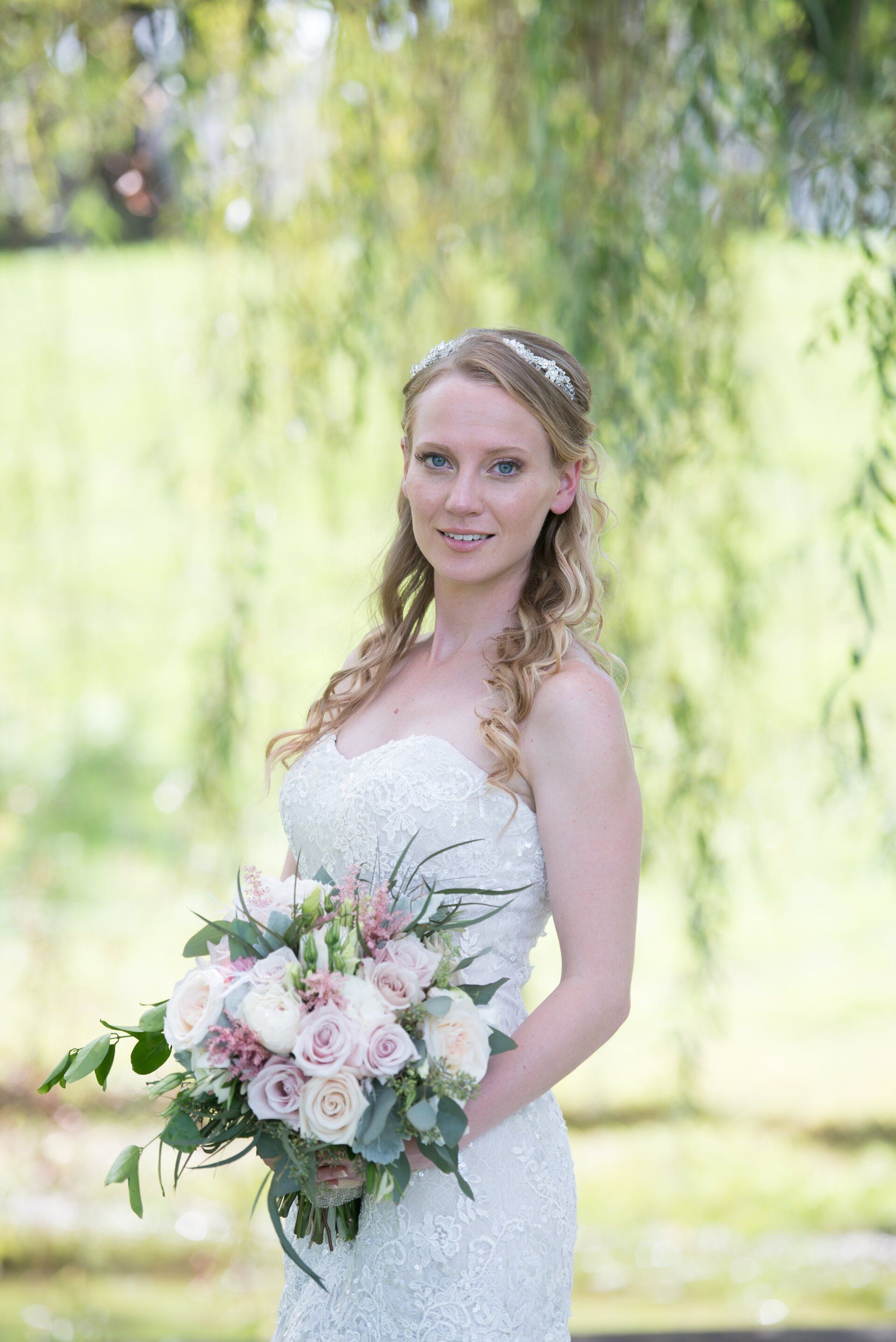 Bride in a white lace gown holding a pastel bouquet outdoors in soft greenery