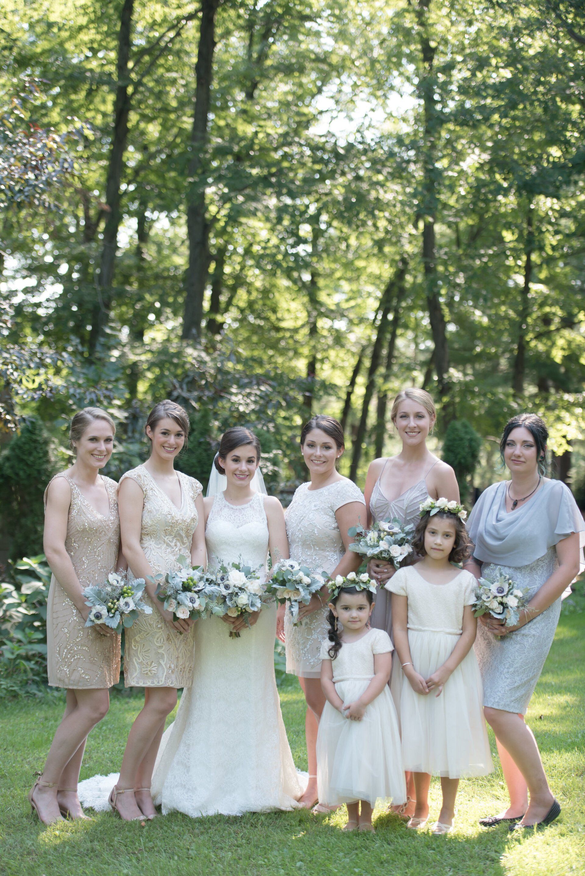 Bridal party in pale dresses and suits posing in a sunlit forest with bouquets