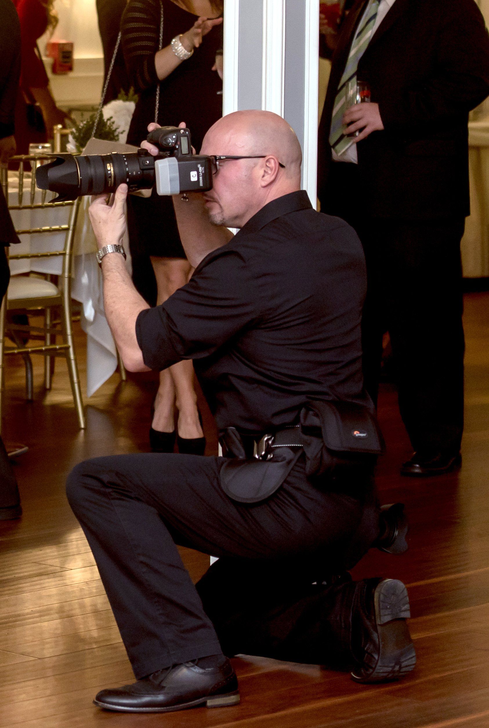 Man kneels on one knee using a DSLR camera with telephoto lens at an indoor event.