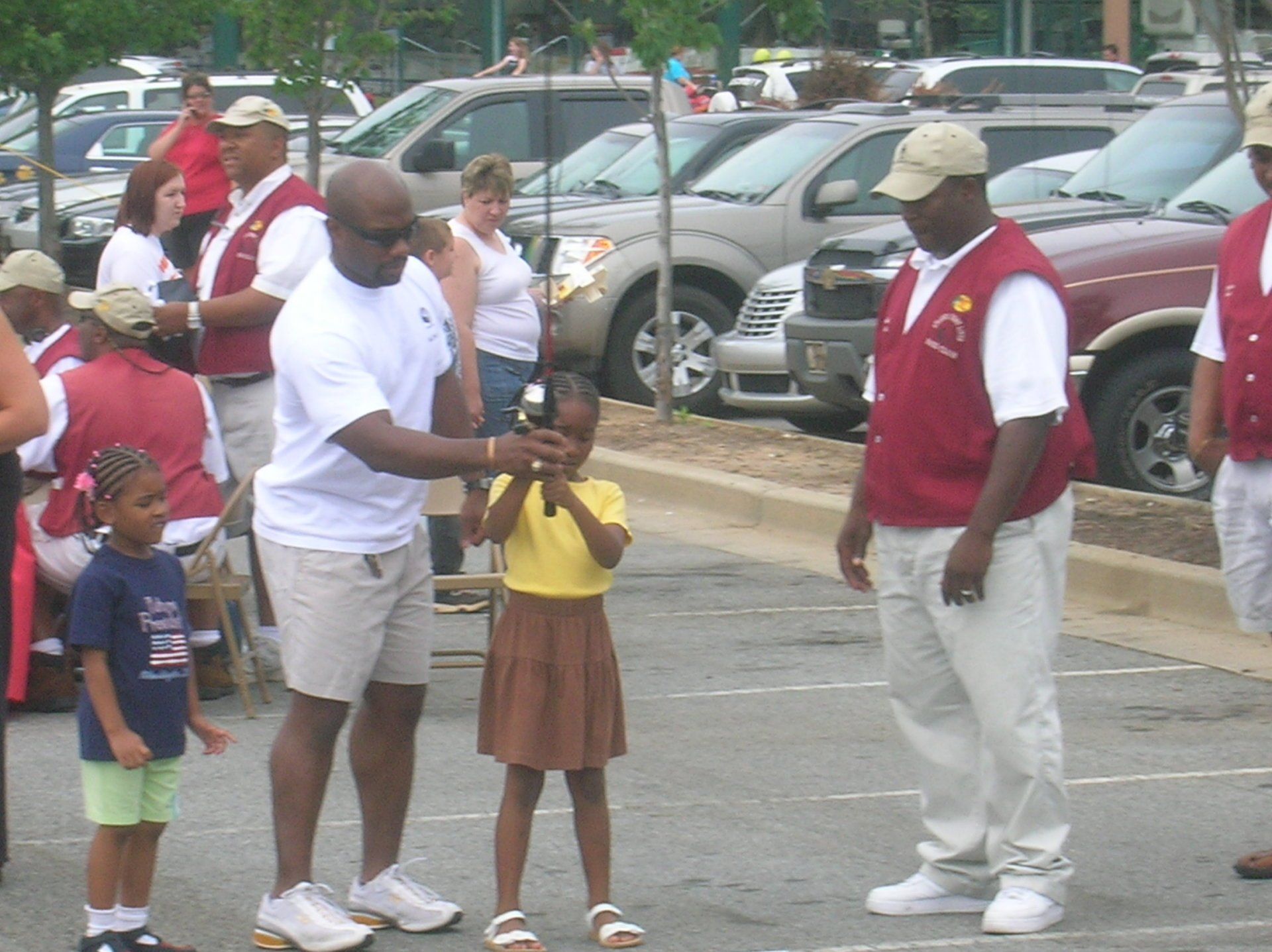 Man helps girl hold a trophy at an outdoor event, other people and cars in background.