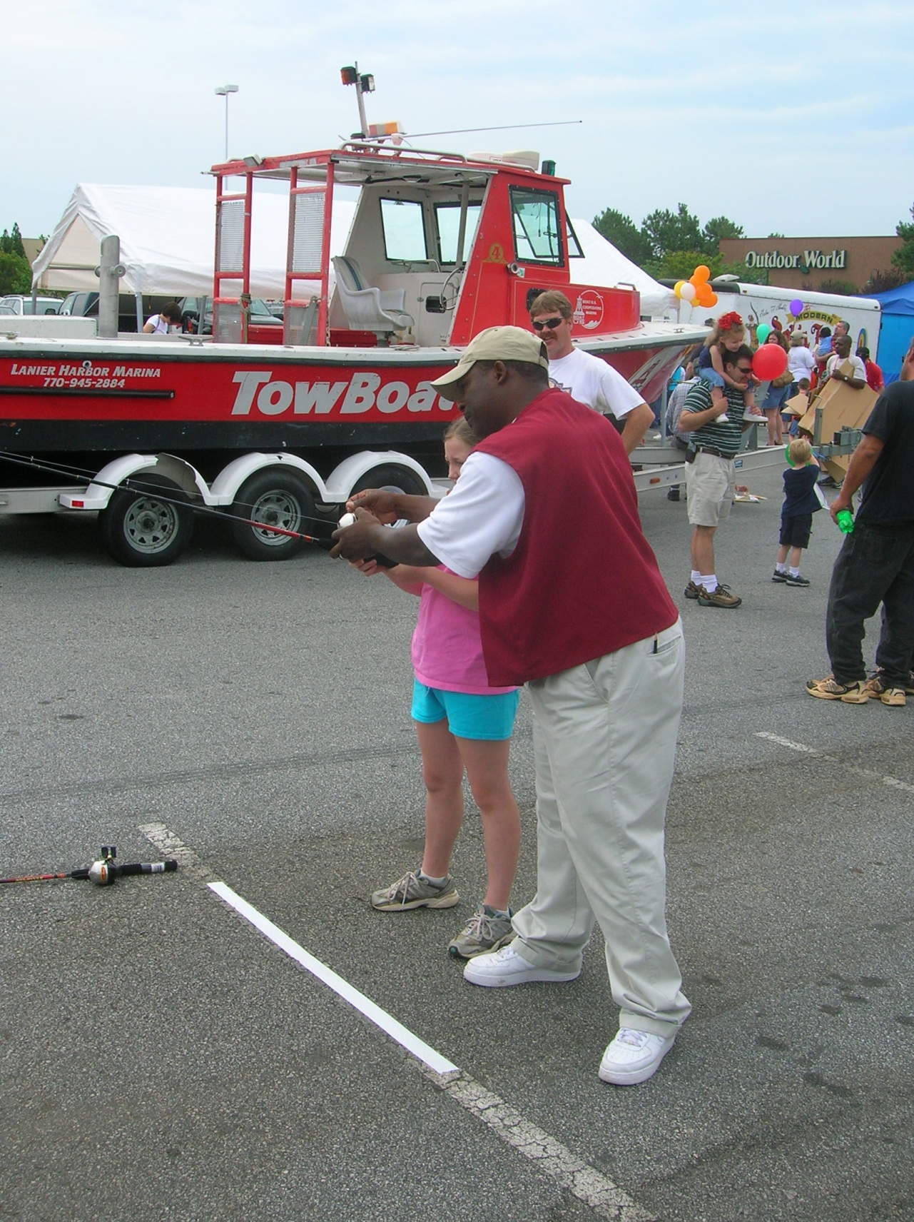 Man teaching a girl to cast a fishing rod in a parking lot, with a red boat in the background.