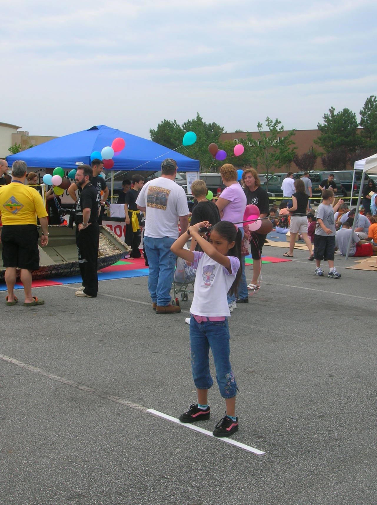 Girl taking a photo at an outdoor event; people, balloons, and tents in the background.