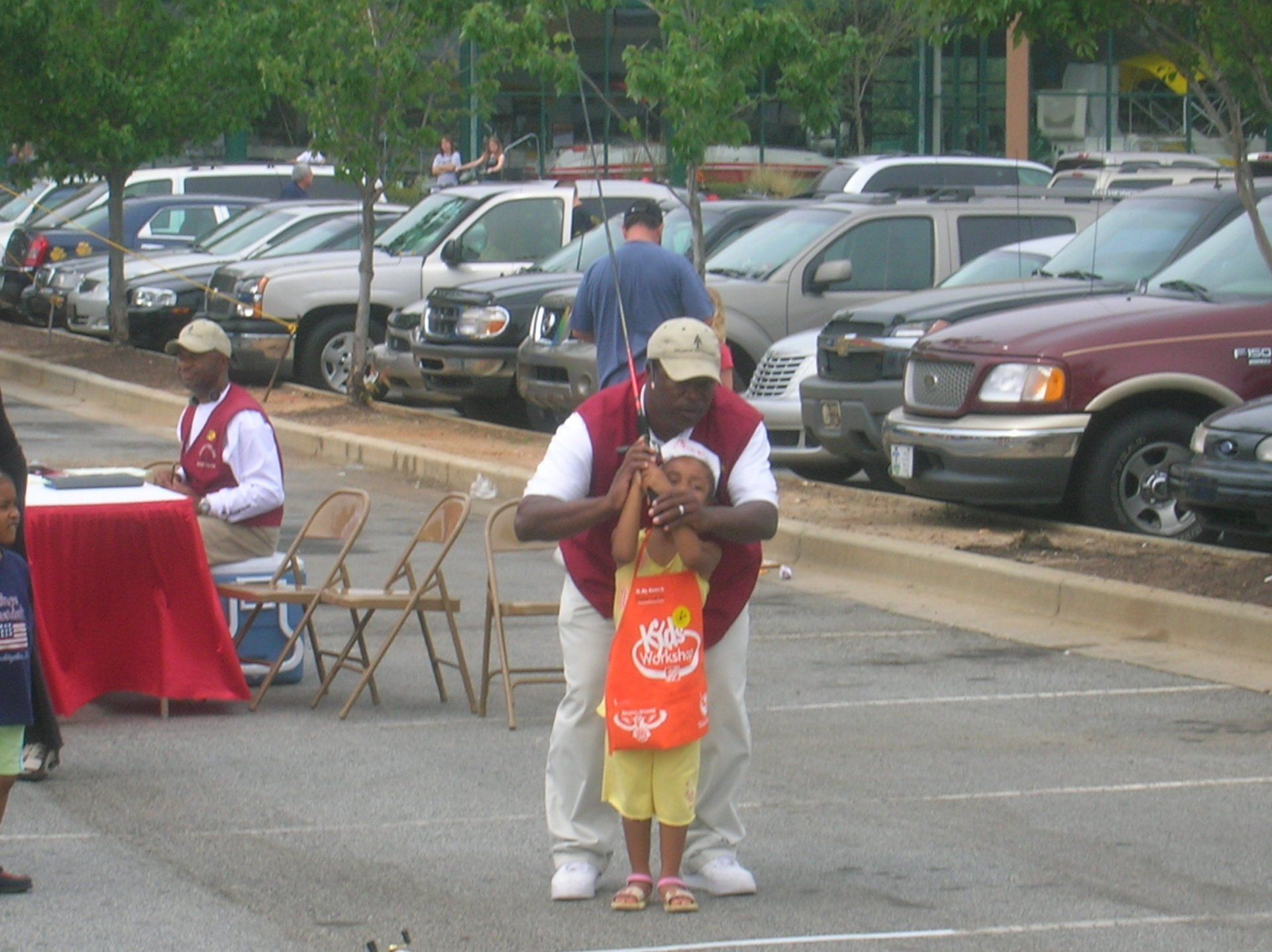 Man holding a small girl in a parking lot. Man is wearing a red vest, and the girl is wearing a bib. Cars in background.