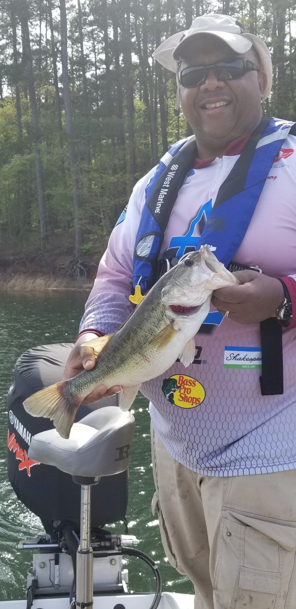 Man on a boat smiles, holding a large fish. Lake in background.