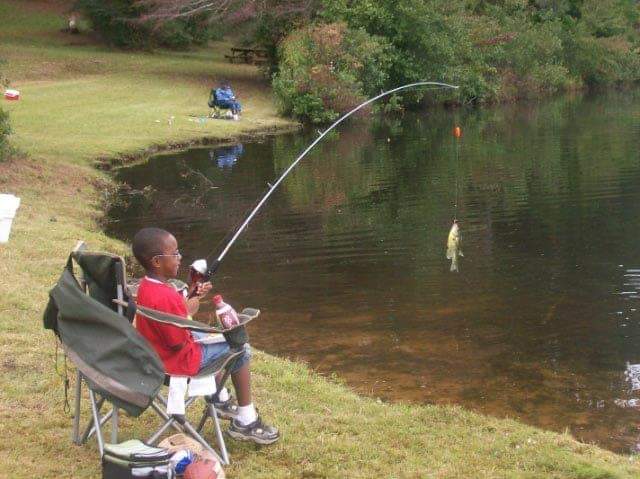 Boy in a red shirt fishing by a lake, sitting in a chair, holding a curved fishing rod.
