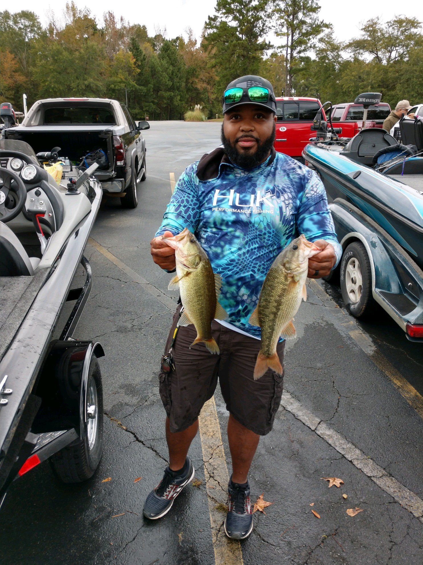 Man holding two bass fish, standing near boats and a truck. He wears a Huk shirt and shorts.