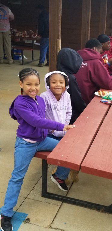 Two smiling Black girls at a picnic table; one on a skateboard, purple hoodie, jeans. Others in the background.
