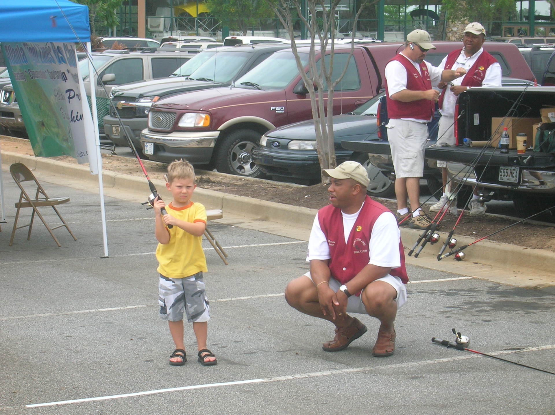 Boy and man fishing at outdoor event, in parking lot. Man kneels, guiding boy holding fishing rod.