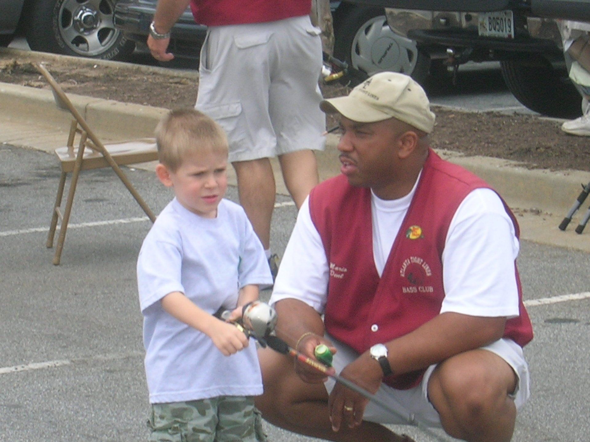 A man in a red vest coaches a young boy holding a fishing rod outdoors.