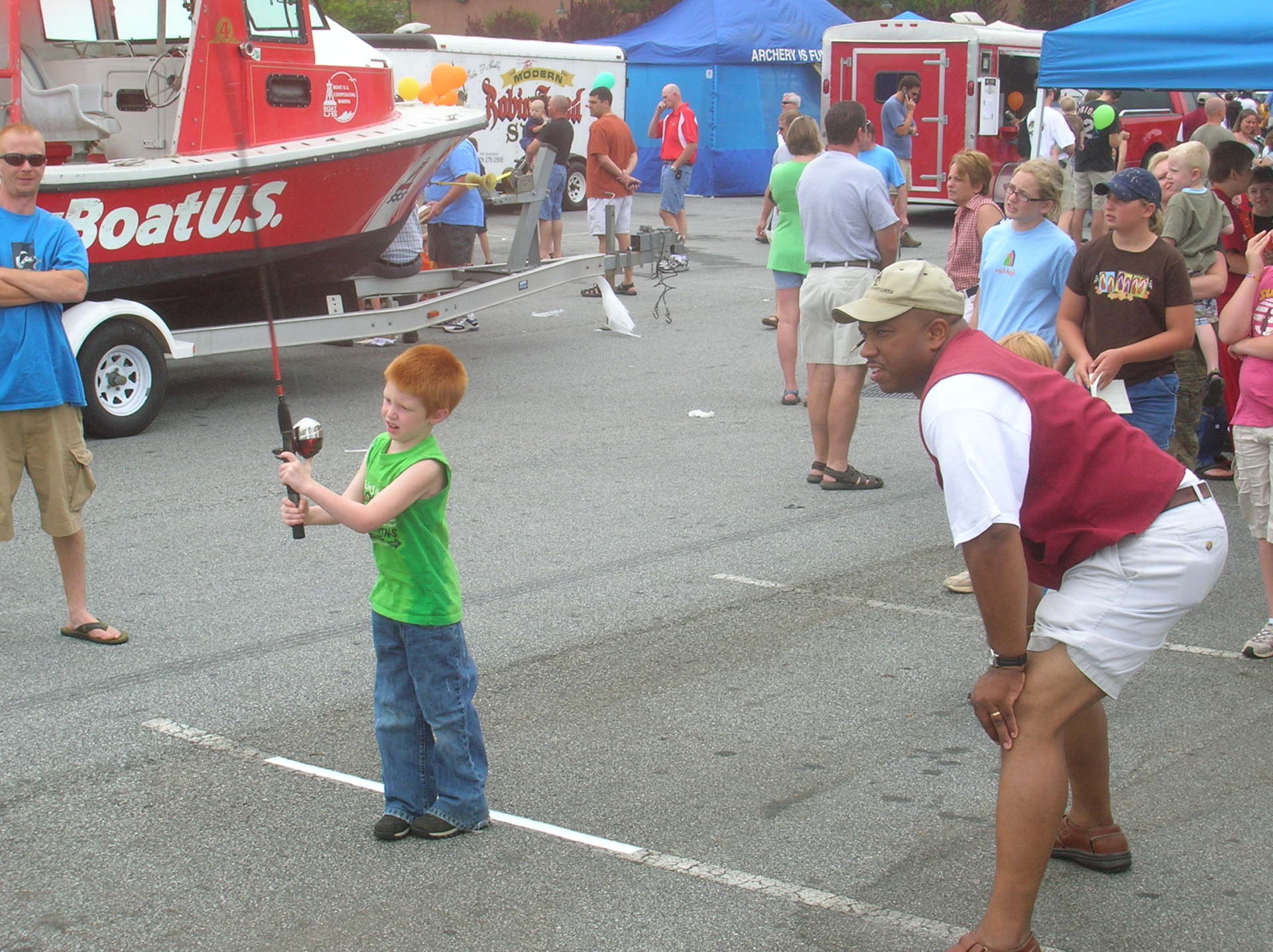 Young boy fishing with an instructor at an outdoor event, red boat in background.