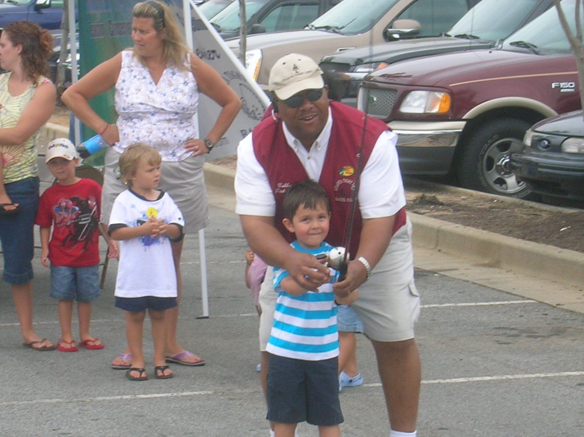 Man assisting a young boy with a camera outside; other people and cars are visible.