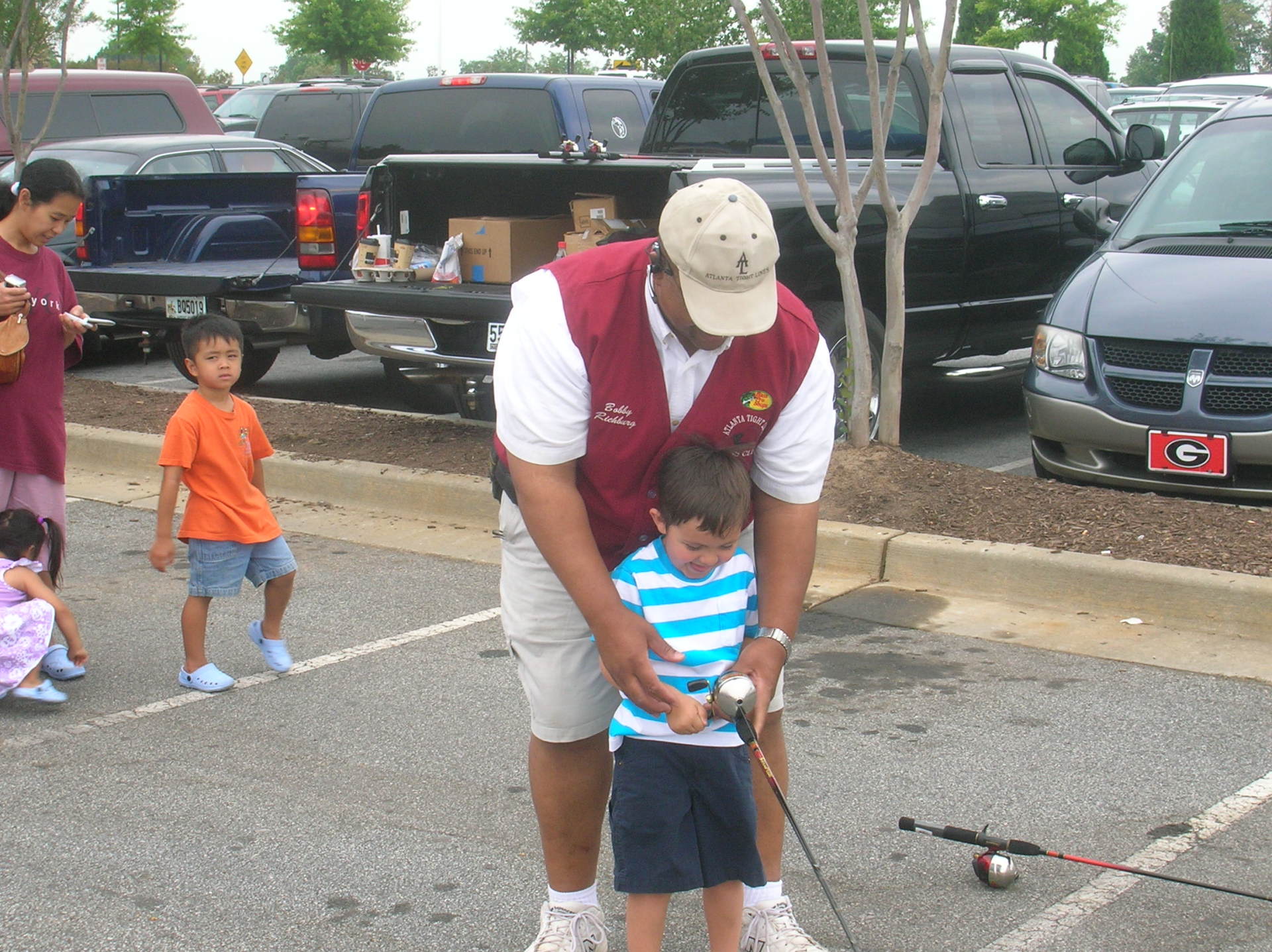 Man teaching a young boy to fish in a parking lot. Other children and vehicles are nearby.