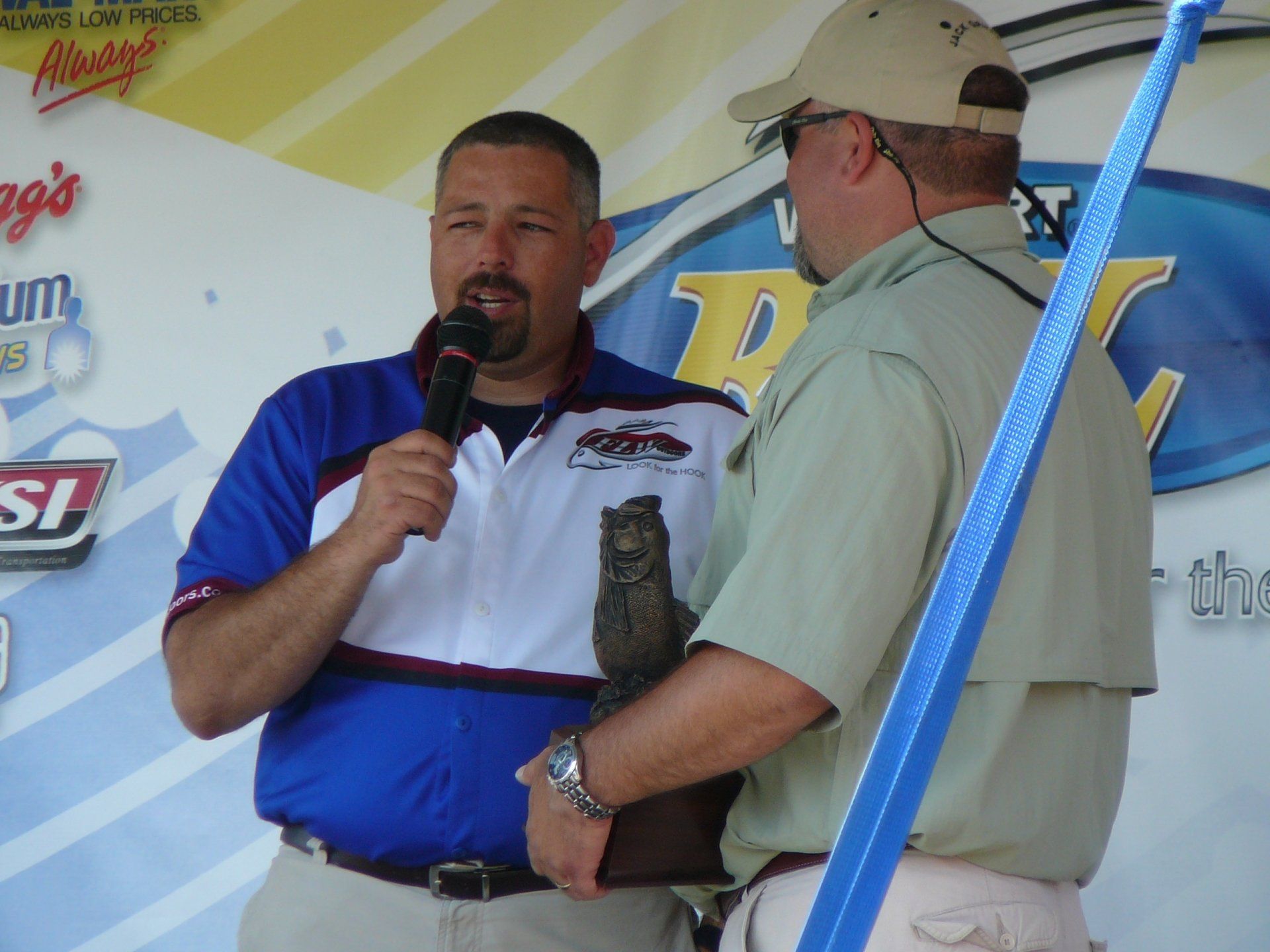 Man in blue shirt speaks, awards trophy to another man. Outdoors, sunny.
