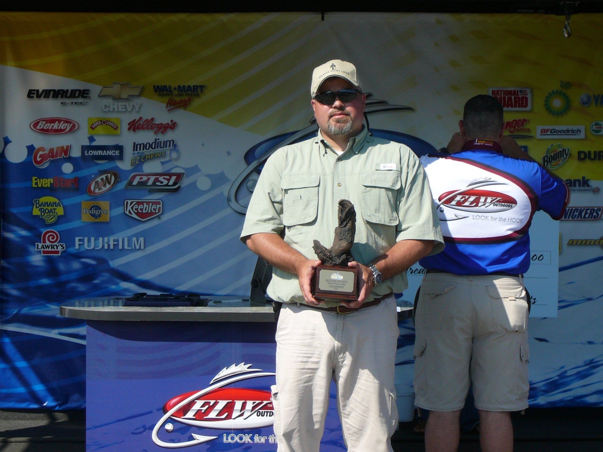 Man holding trophy, posing at a tournament with a background of logos and another man to his right.